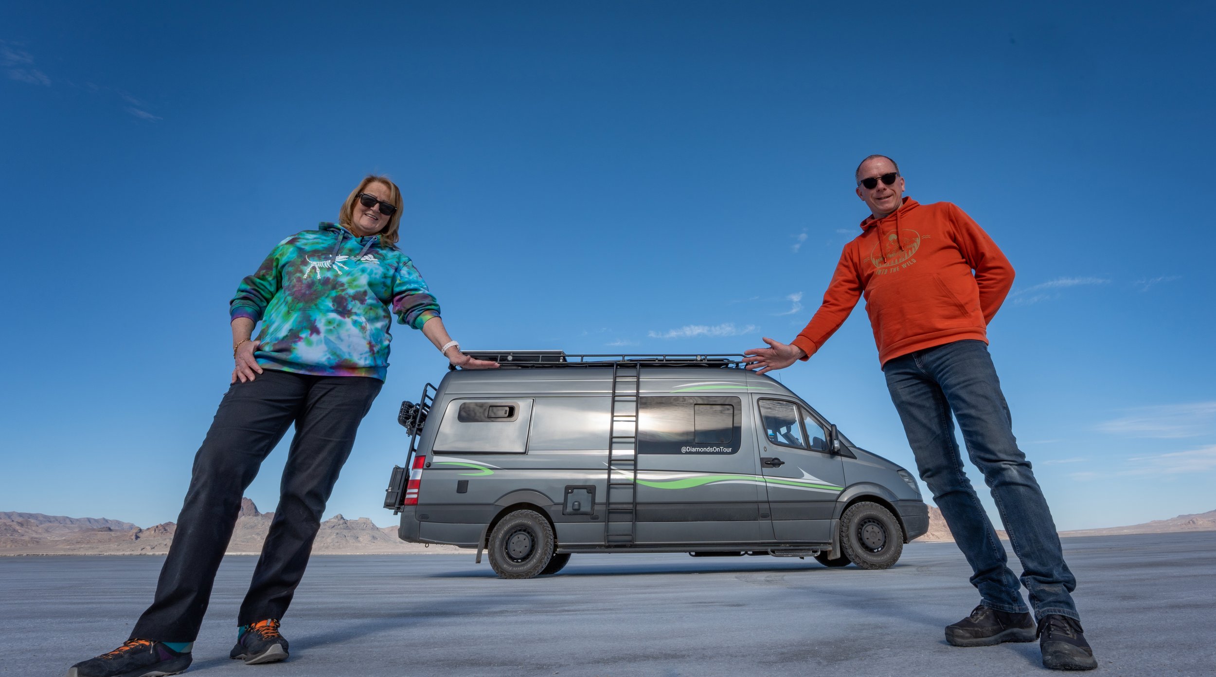 Two people, a woman in a colorful hoodie and a man in an orange hoodie, standing on a flat, open landscape with a small van between them, under a clear blue sky.