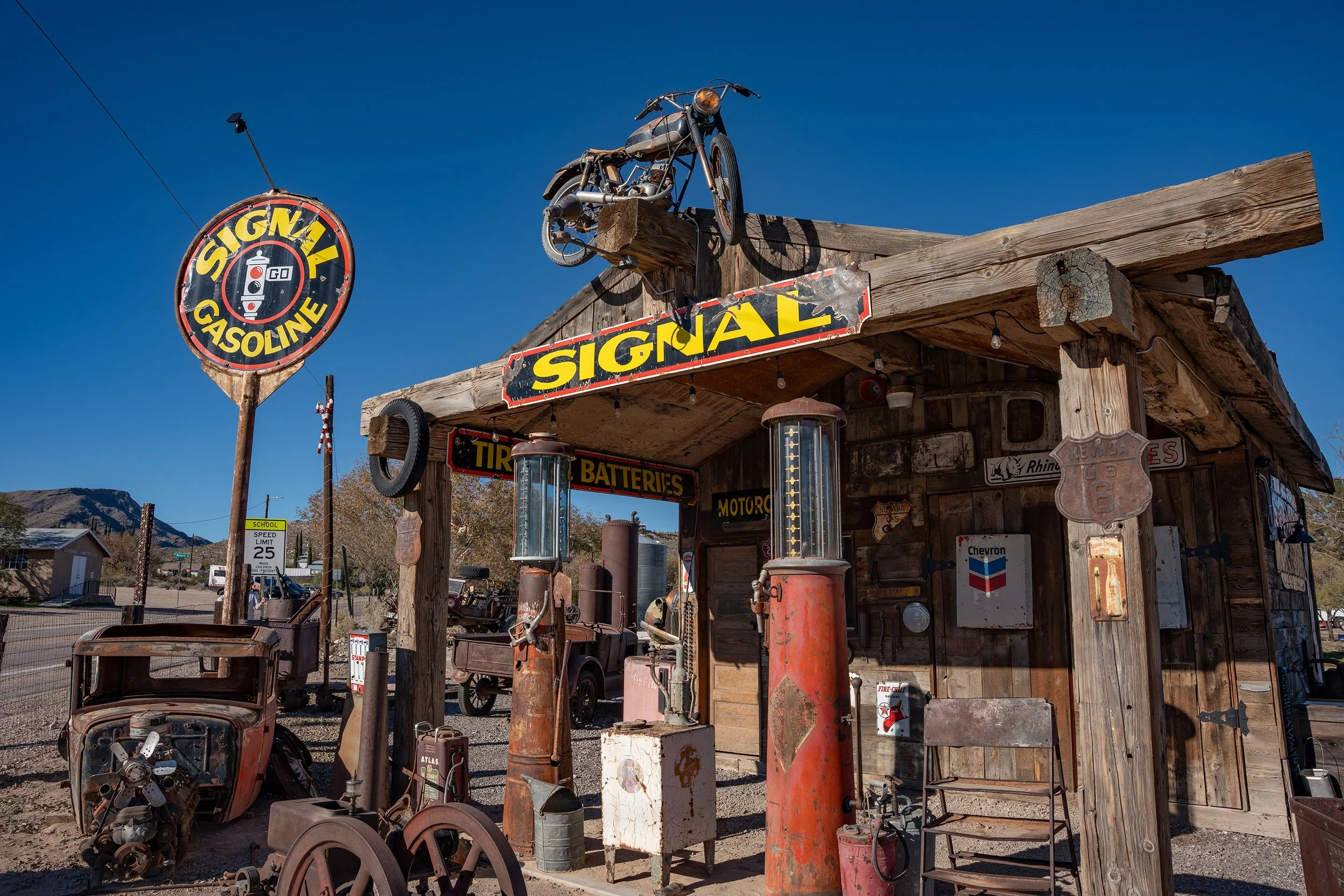 Old gas station with vintage gasoline pumps, rusty car parts, and a sign that reads 'Signal Gasoline,' located in a rural area with mountains in the background.