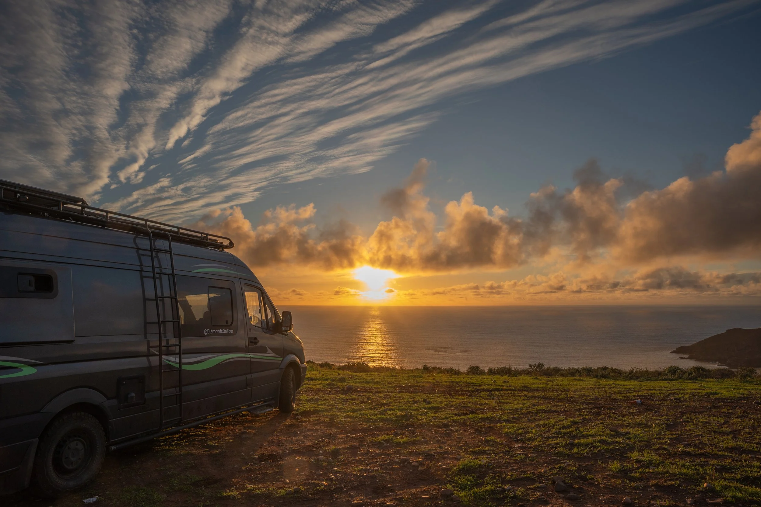 A grey camper van parked on a grassy area overlooking the ocean at sunset, with clouds and a partly cloudy sky.