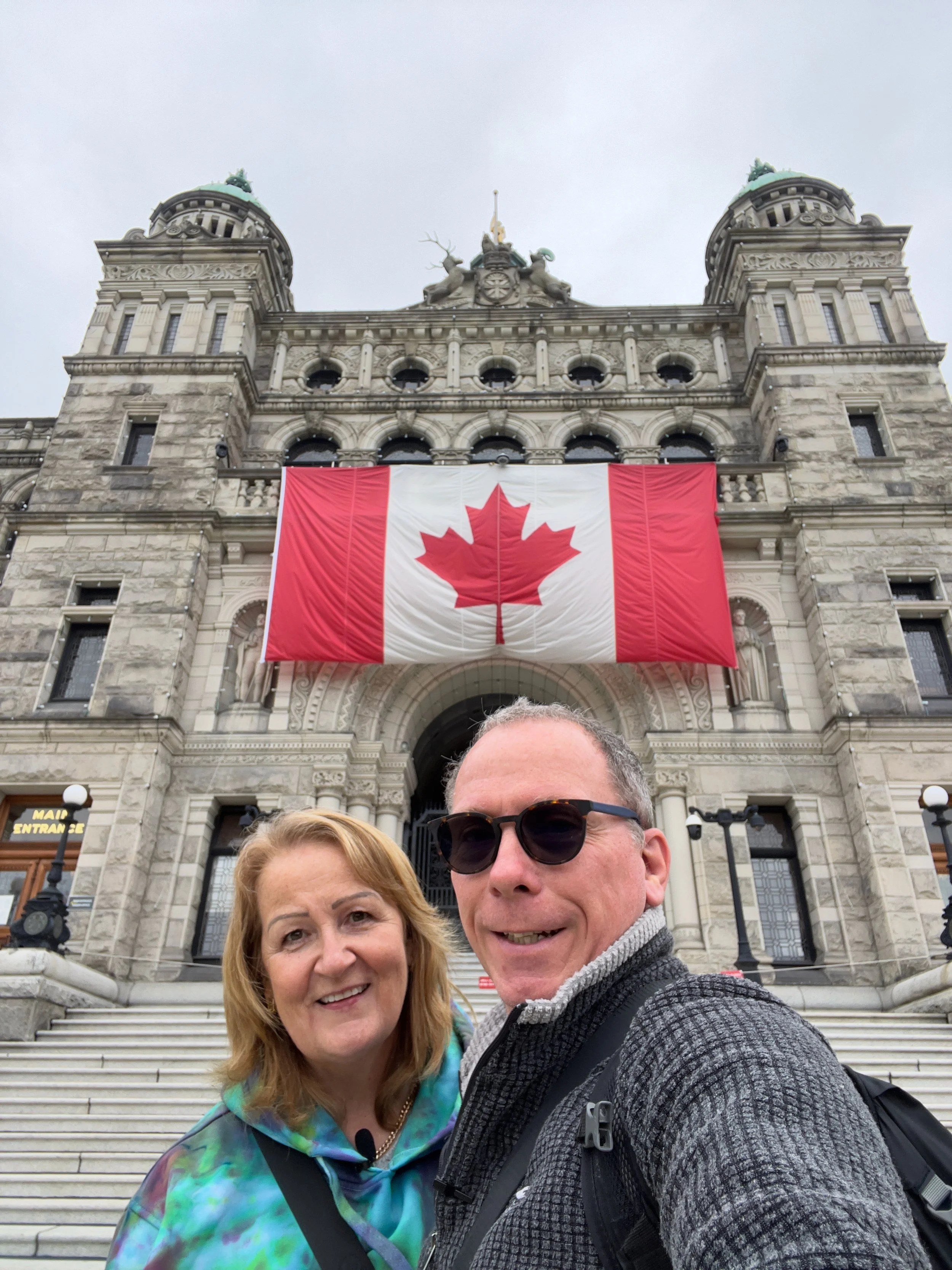 A smiling woman and man taking a selfie in front of a historic stone building with stairs, large banner of the Canadian flag, and architectural details like towers, pillars, and sculptures.