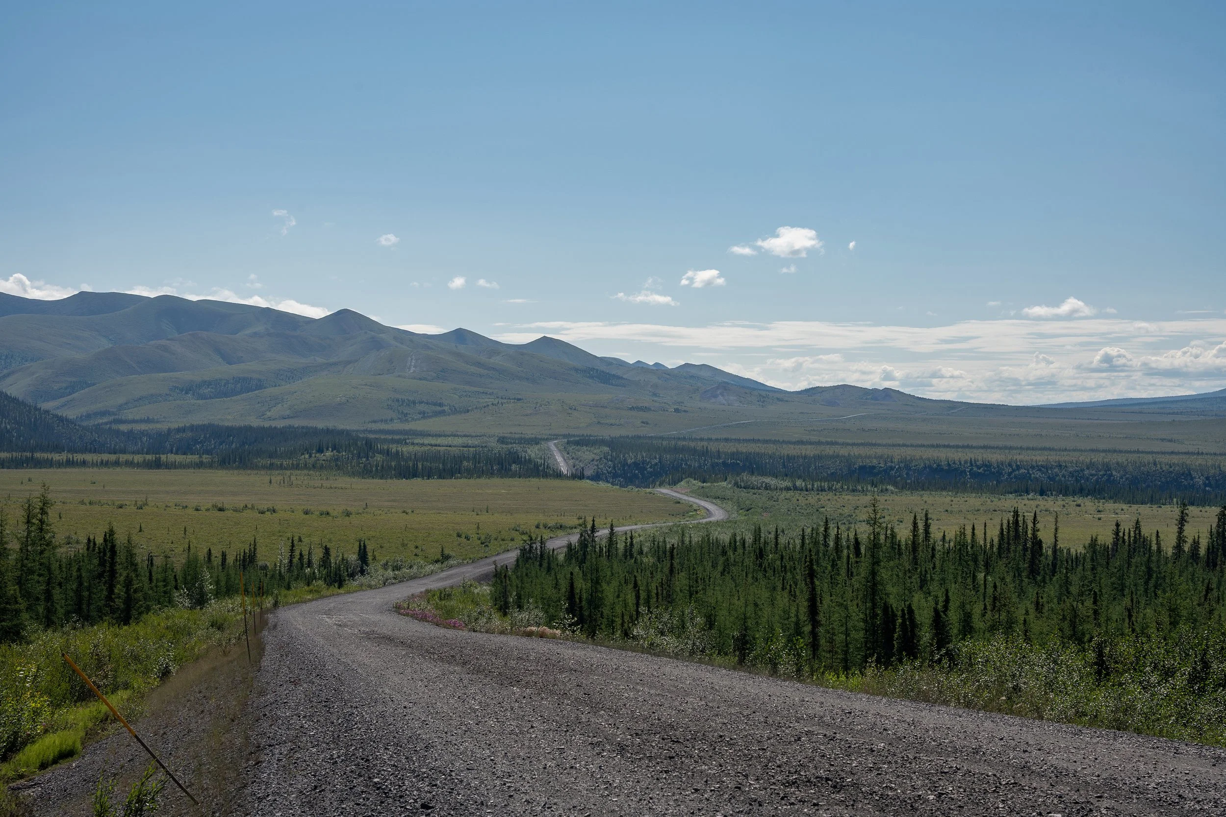 A gravel road winding through a vast green forest with mountains in the background under a partly cloudy blue sky.
