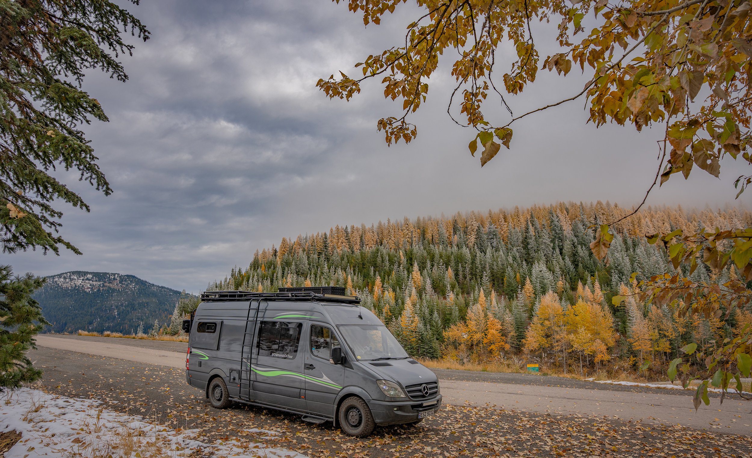A gray camper van parked on the side of a mountain road surrounded by autumn trees with orange, yellow, and green leaves, some snow on the ground, with mountains and a cloudy sky in the background.
