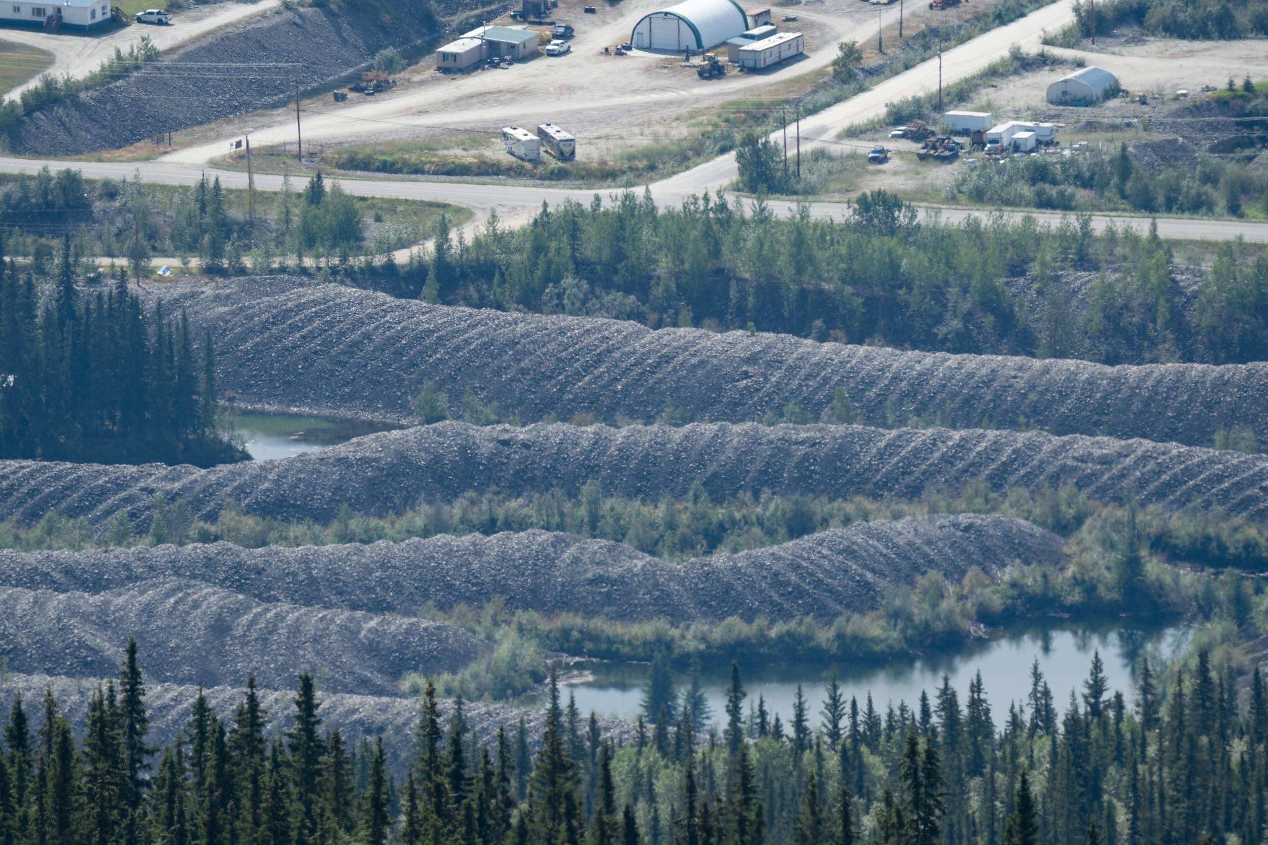 Aerial view of a rural industrial area with storage buildings, parked vehicles, dirt roads, and surrounding forested landscape. Gold mining tailings, Dawson City, Yukon Territory, Canada.