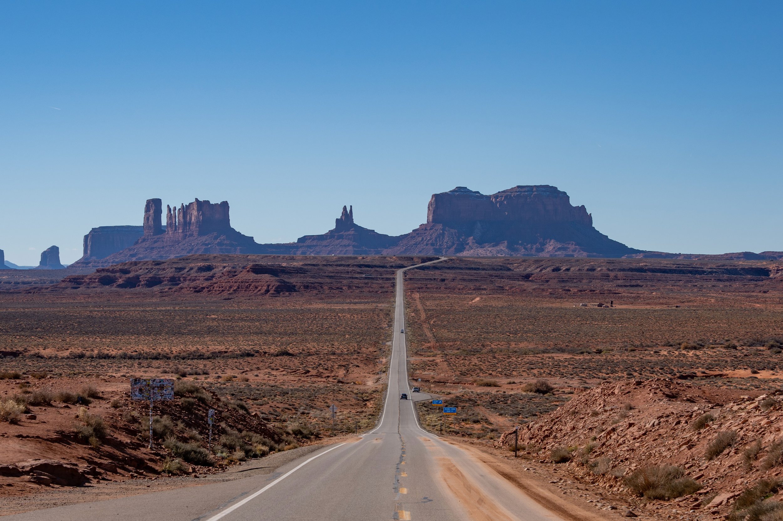 A straight road extending through a desert landscape with large rock formations in the distance under a clear blue sky. Monument Valley Forrest Gump Point.