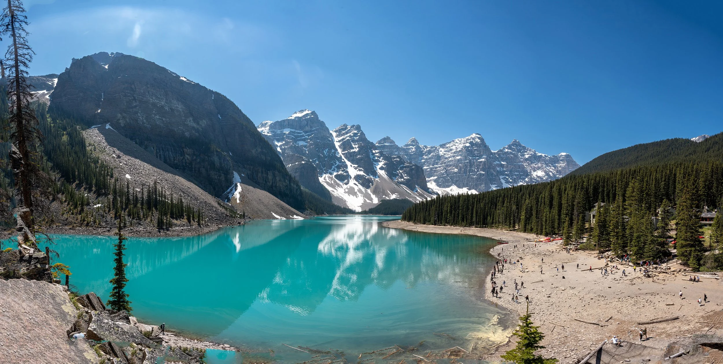 A scenic view of a large turquoise lake surrounded by pine trees and snow-capped mountains under a clear blue sky. Moraine Lake.