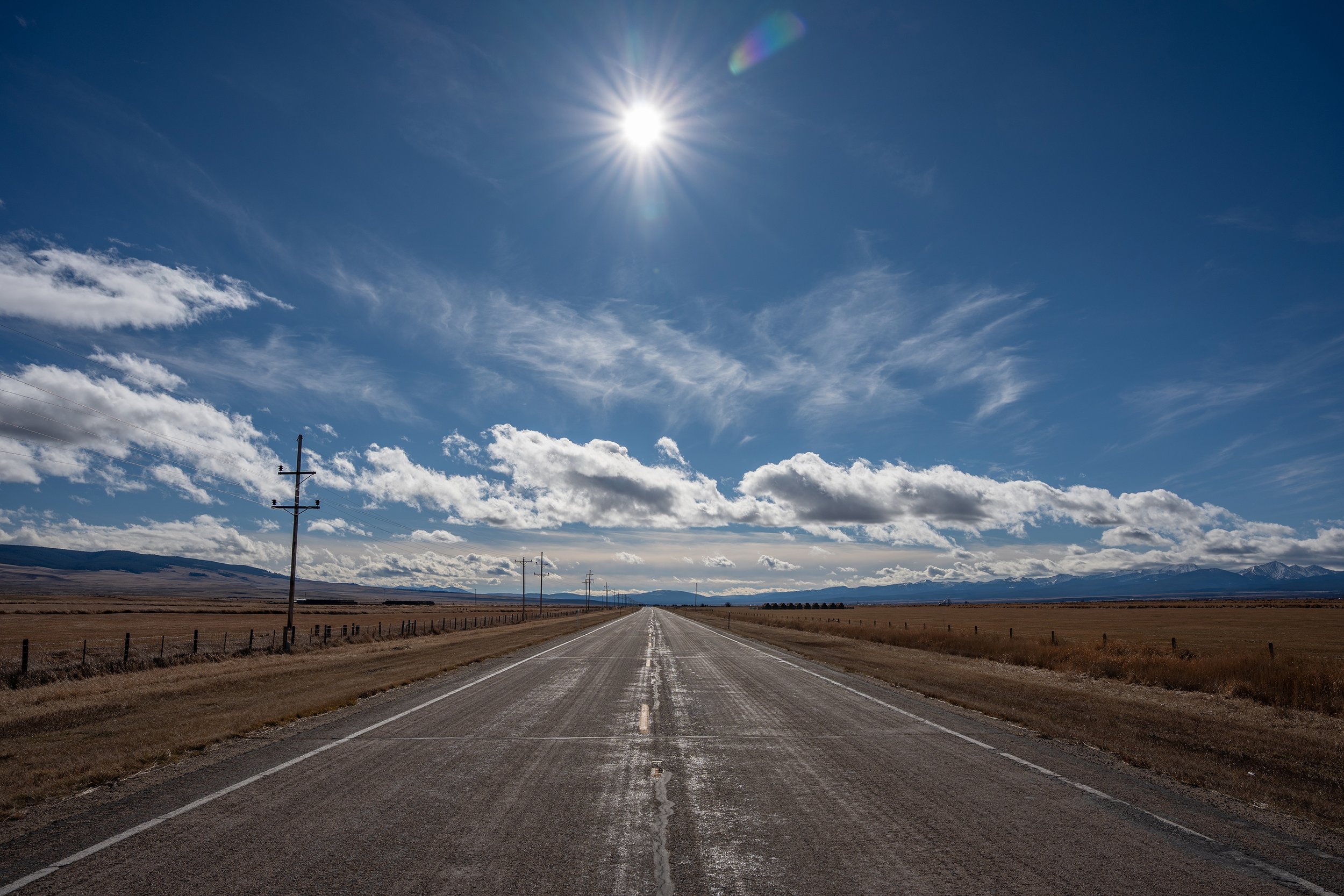 Long straight road stretching into the distance through an open landscape with mountains in the background, a blue sky with scattered clouds, and the sun shining brightly overhead.