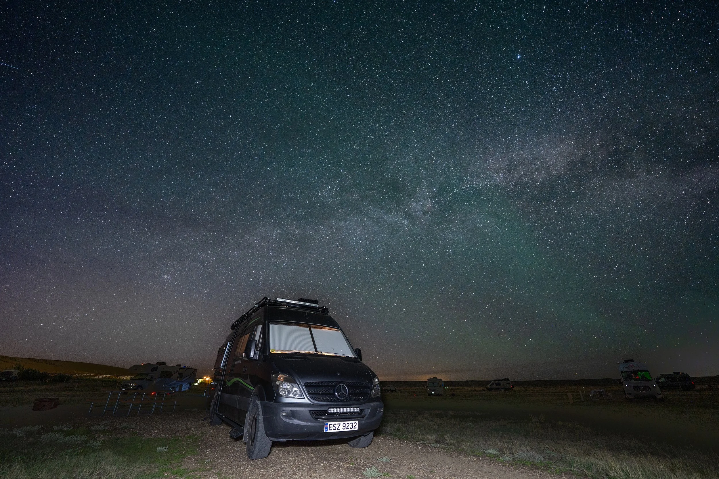 Night sky filled with stars and the Milky Way galaxy visible above a camper van with a roof tent parked in a campground.