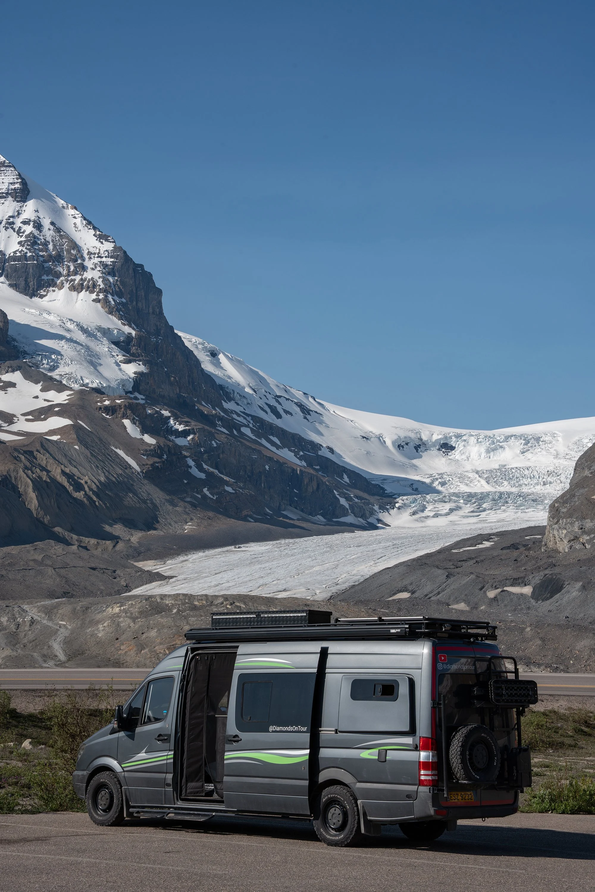 A camper van parked on the side of a mountain road with snow-capped mountains in the background and a clear blue sky above. Athabasca Glacier, Canada.