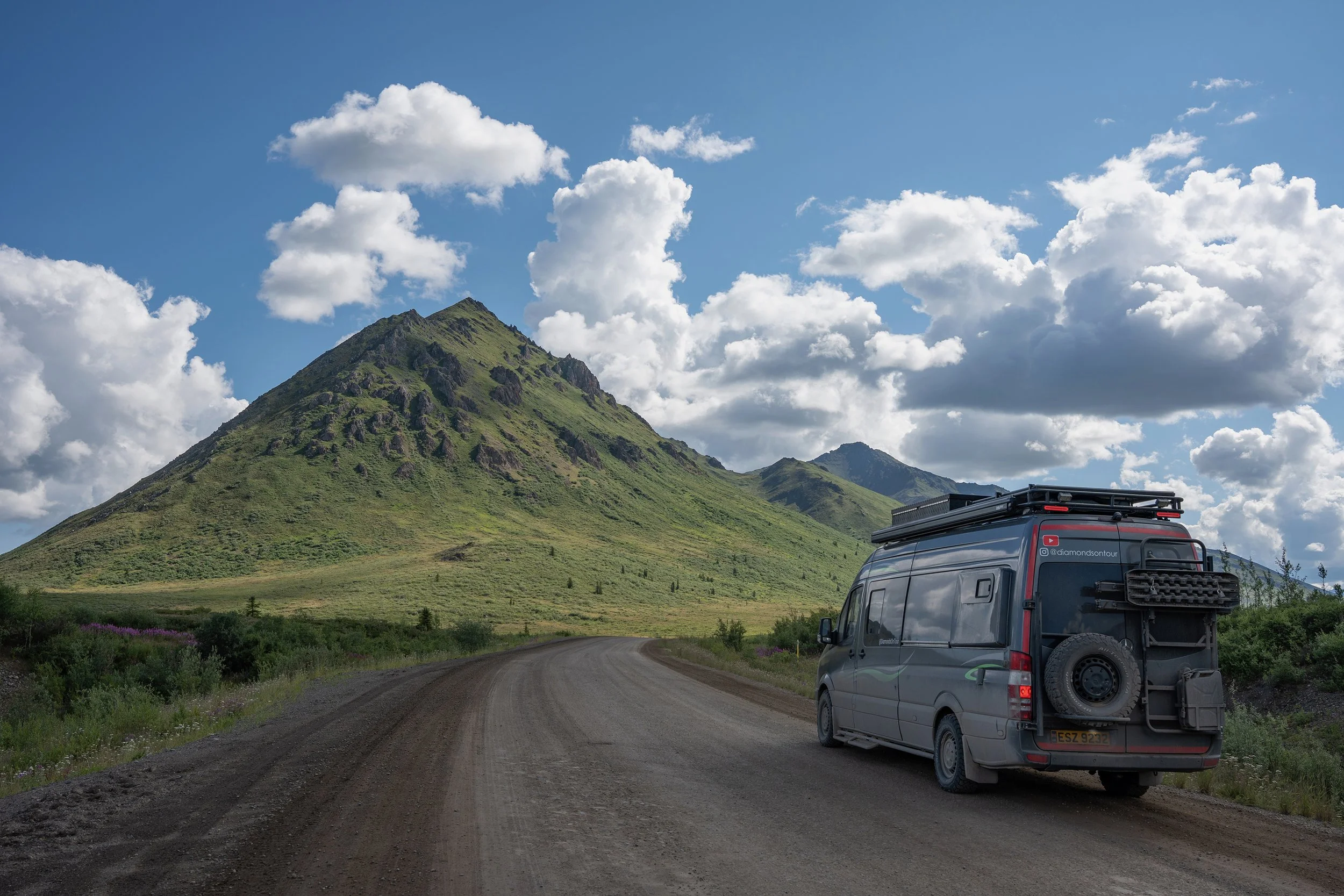 A scenic landscape with a mountain covered in green vegetation under a partly cloudy sky. In the foreground, a gravel road and a parked dark gray van with a roof rack are visible.