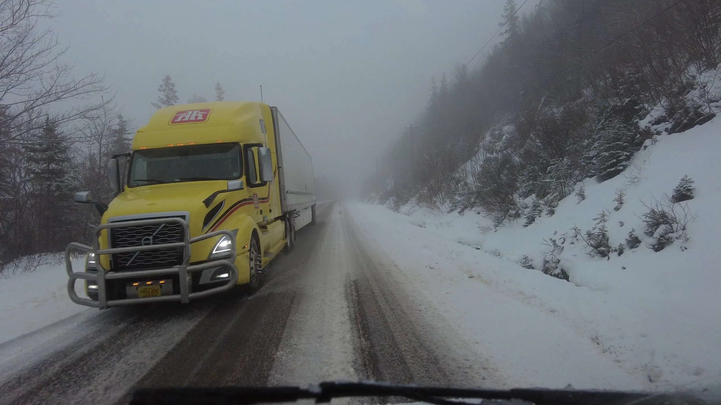 Yellow semi-truck driving on a snow-covered mountain road with foggy weather and snow on the trees and hillside.