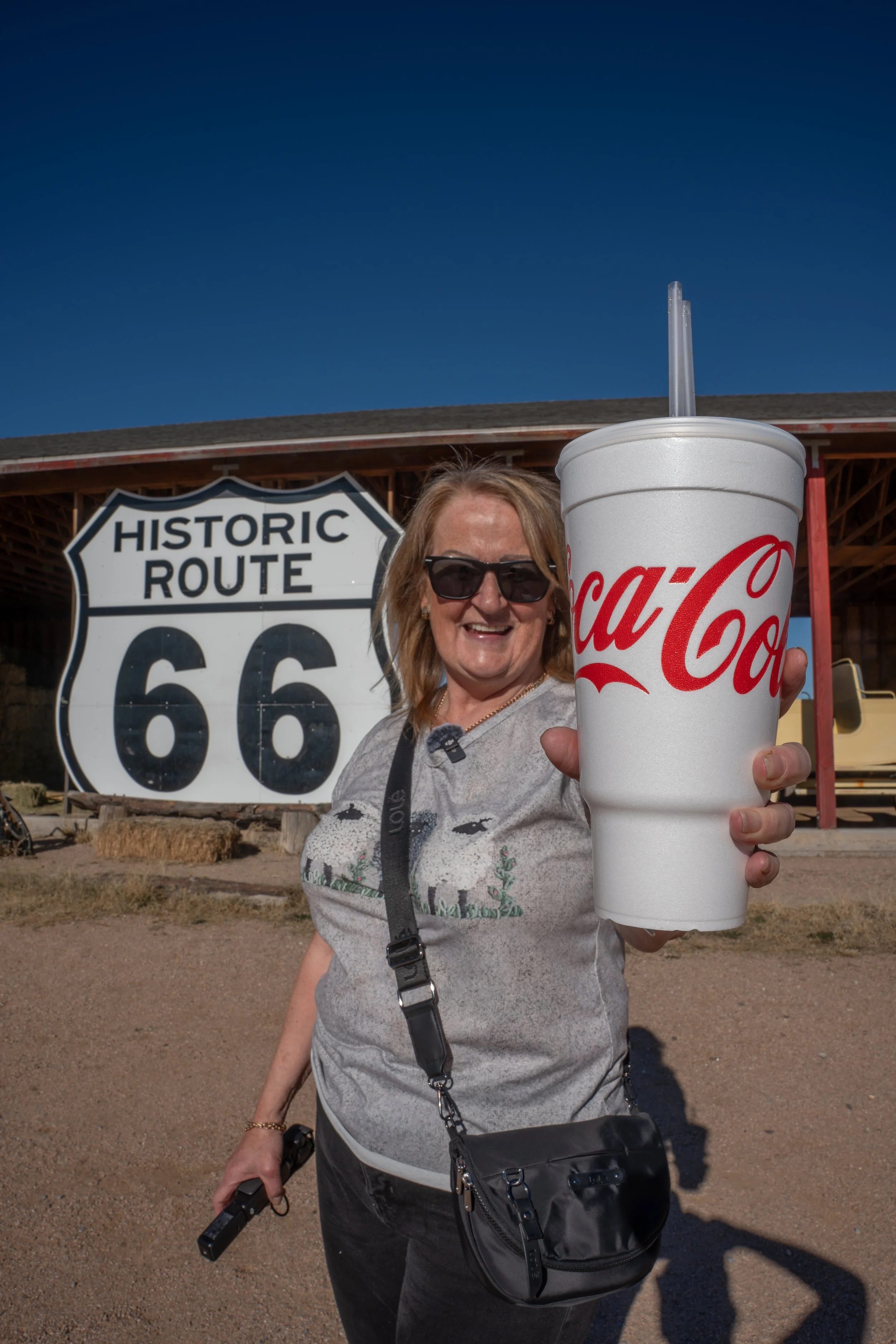 A woman wearing sunglasses smiling while holding a large Coca-Cola cup in front of a Route 66 sign at a historic site.