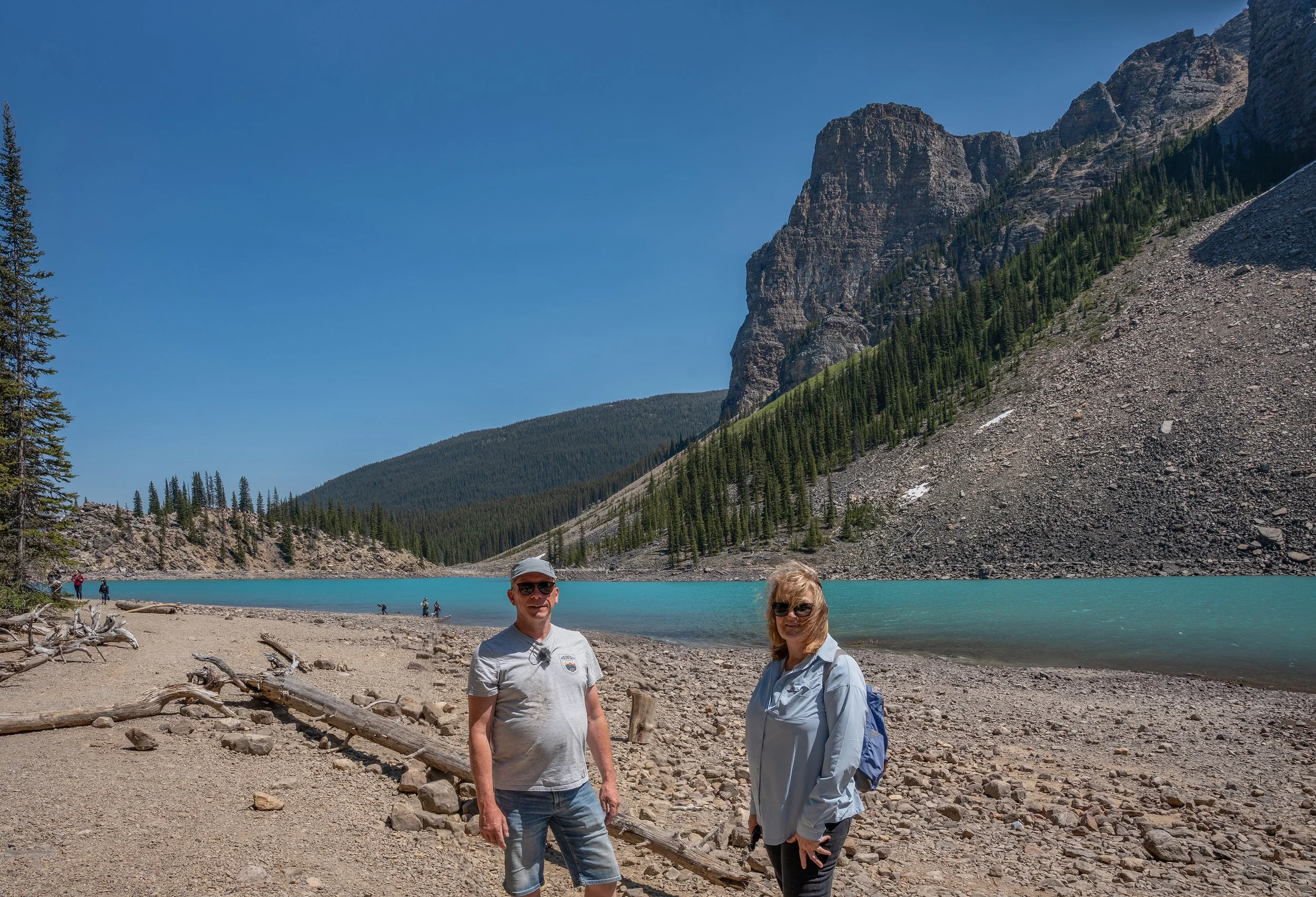 Two people standing on a rocky beach in front of a turquoise lake with mountains and pine trees in the background. The sky is clear and blue. Moraine Lake, Rocky Mountains, Canada.