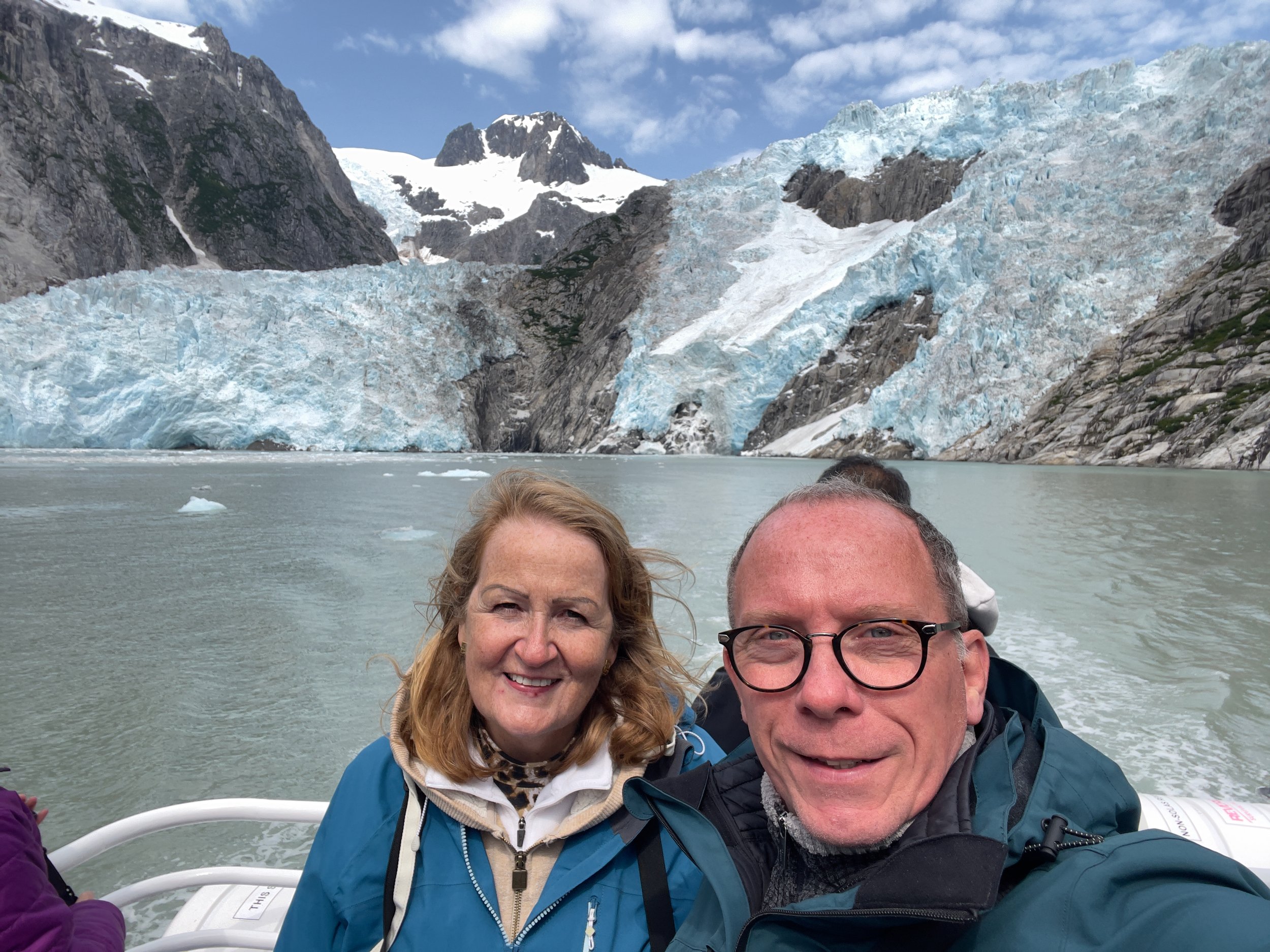 Two people smiling, taking a selfie on a boat with glacier-covered mountains and icy waters in the background. Alaska boat trip from Seward.