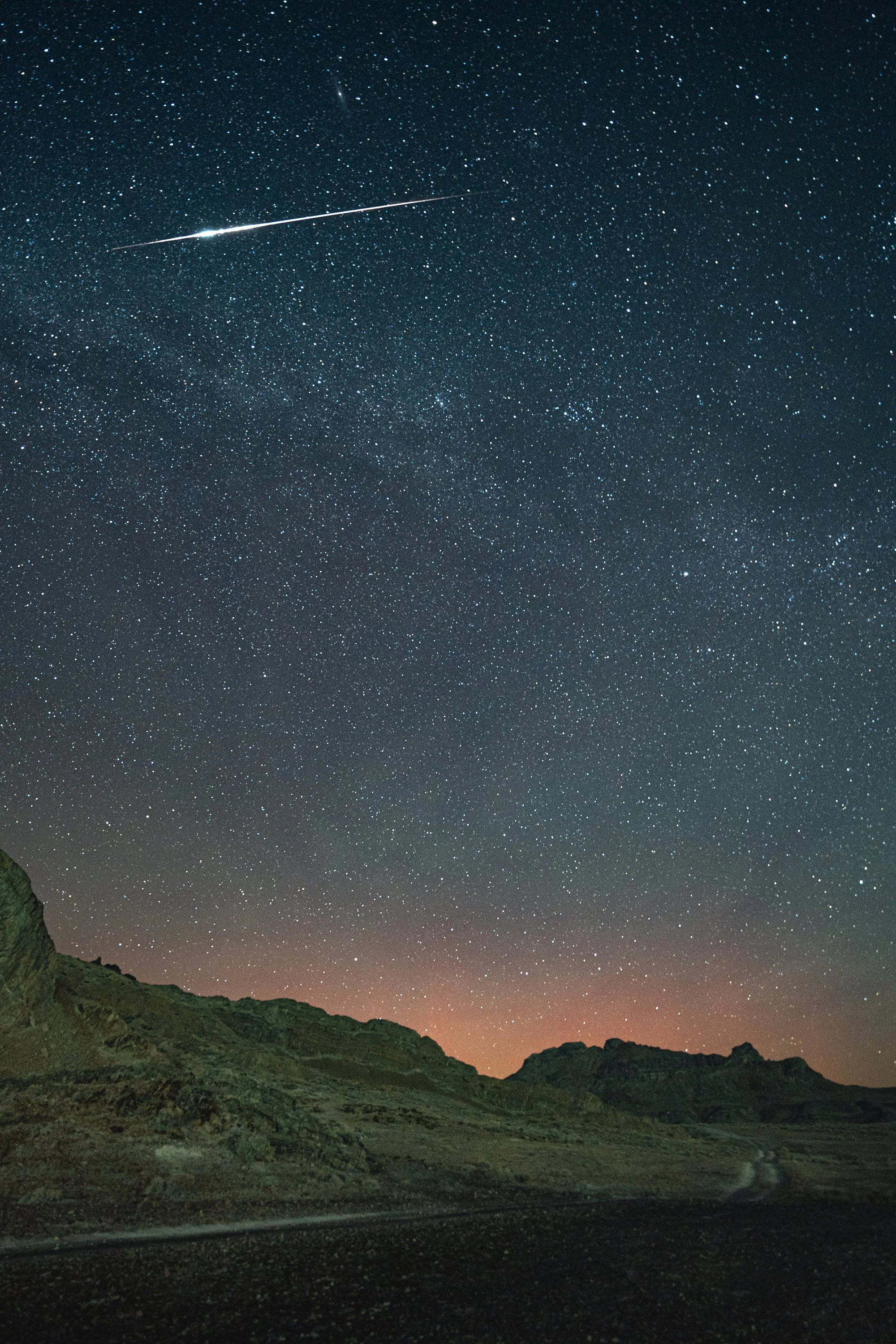 A clear night sky filled with countless stars, with a bright shooting star or meteor streaking across the sky above a mountainous landscape with rocky terrain and some greenery.