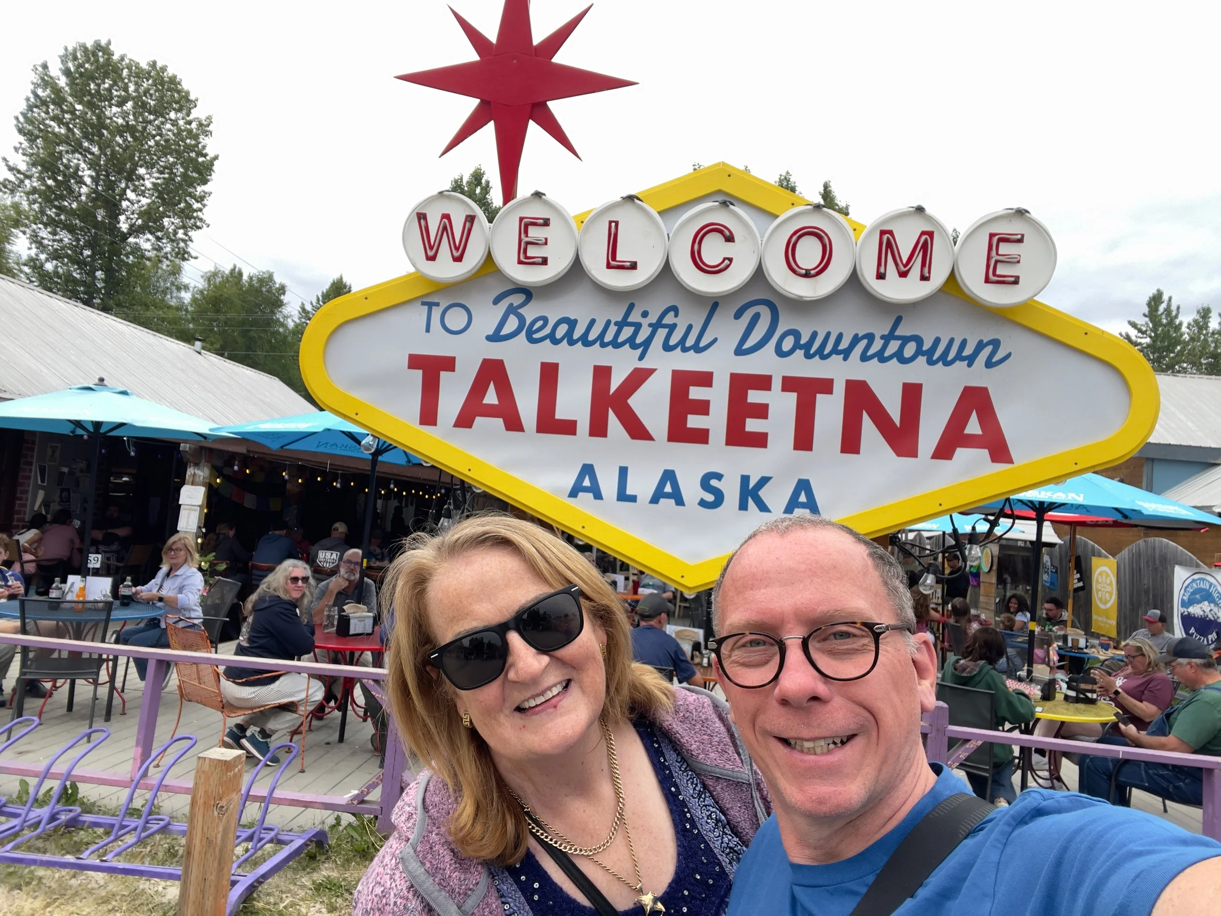 A joyful woman and man taking a selfie in front of a large welcome sign for Downtown Talkeetna, Alaska, with outdoor tables, patrons, and trees in the background on a cloudy day.