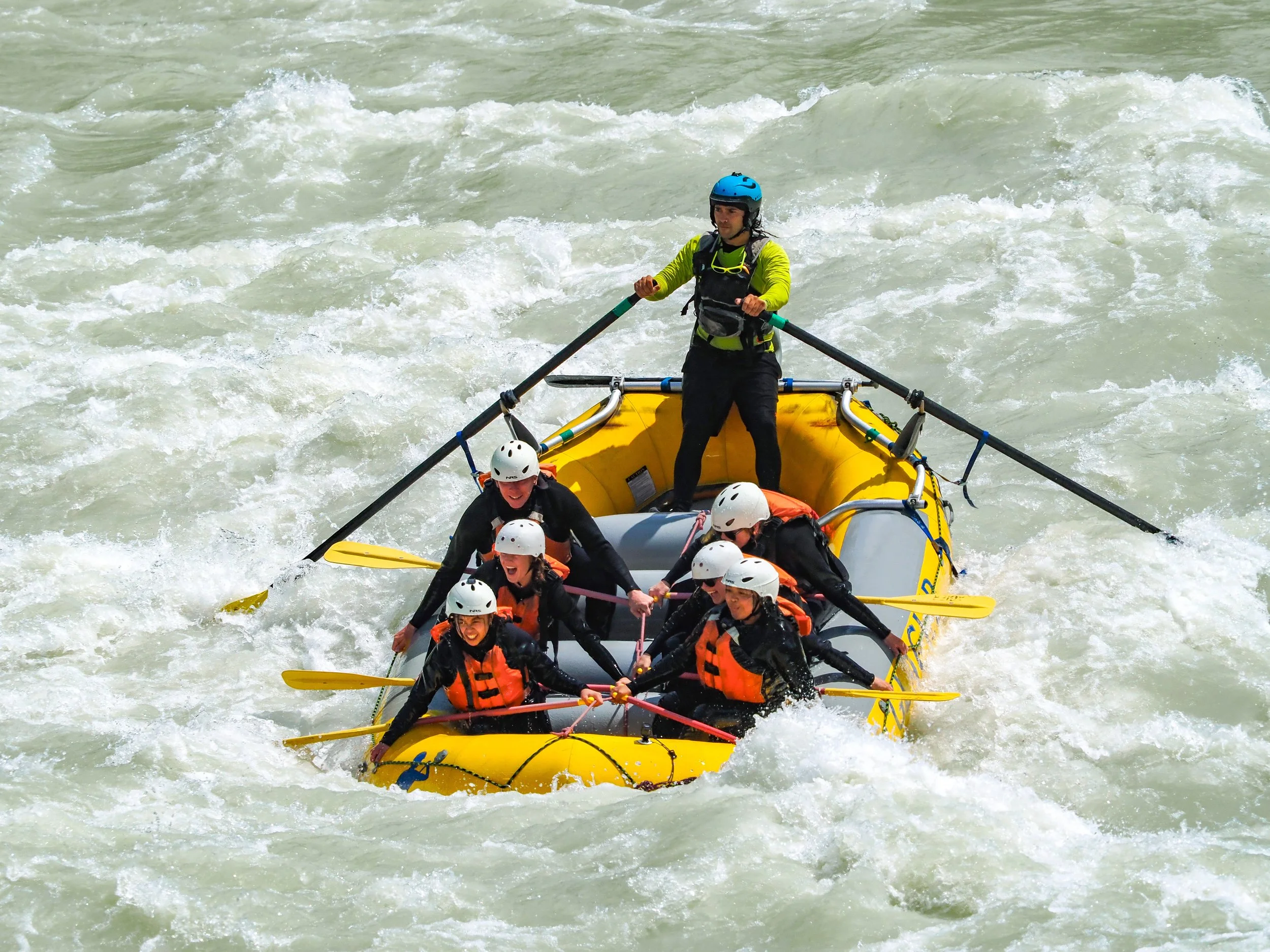 A group of people on a yellow inflatable raft, white helmets, and life jackets navigating white water rapids. One person stands at the front with a helmet and a yellow long-sleeve shirt, while others sit or hold the paddles, all enjoying an outdoor w
