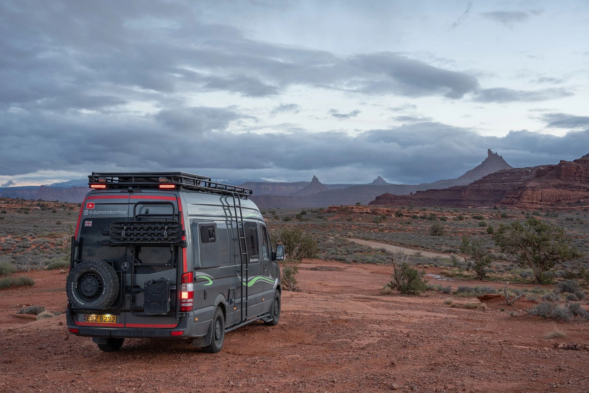 A camper van parked on a dirt road in a desert landscape with red rocks and distant mesas under a cloudy sky.