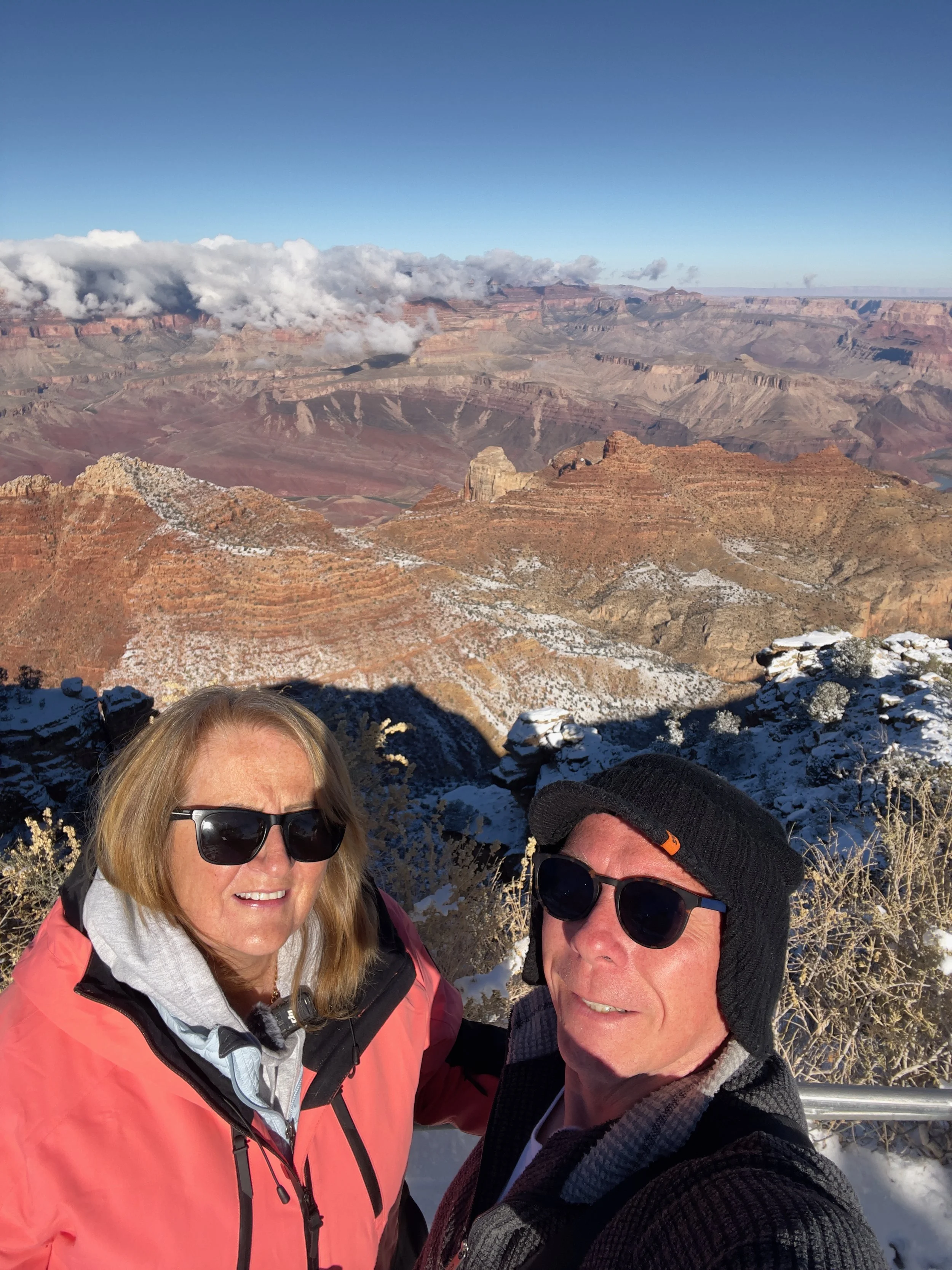 Two people taking a selfie at the Grand Canyon with snowy rocks in the foreground and the canyon's layered rock formations in the background.