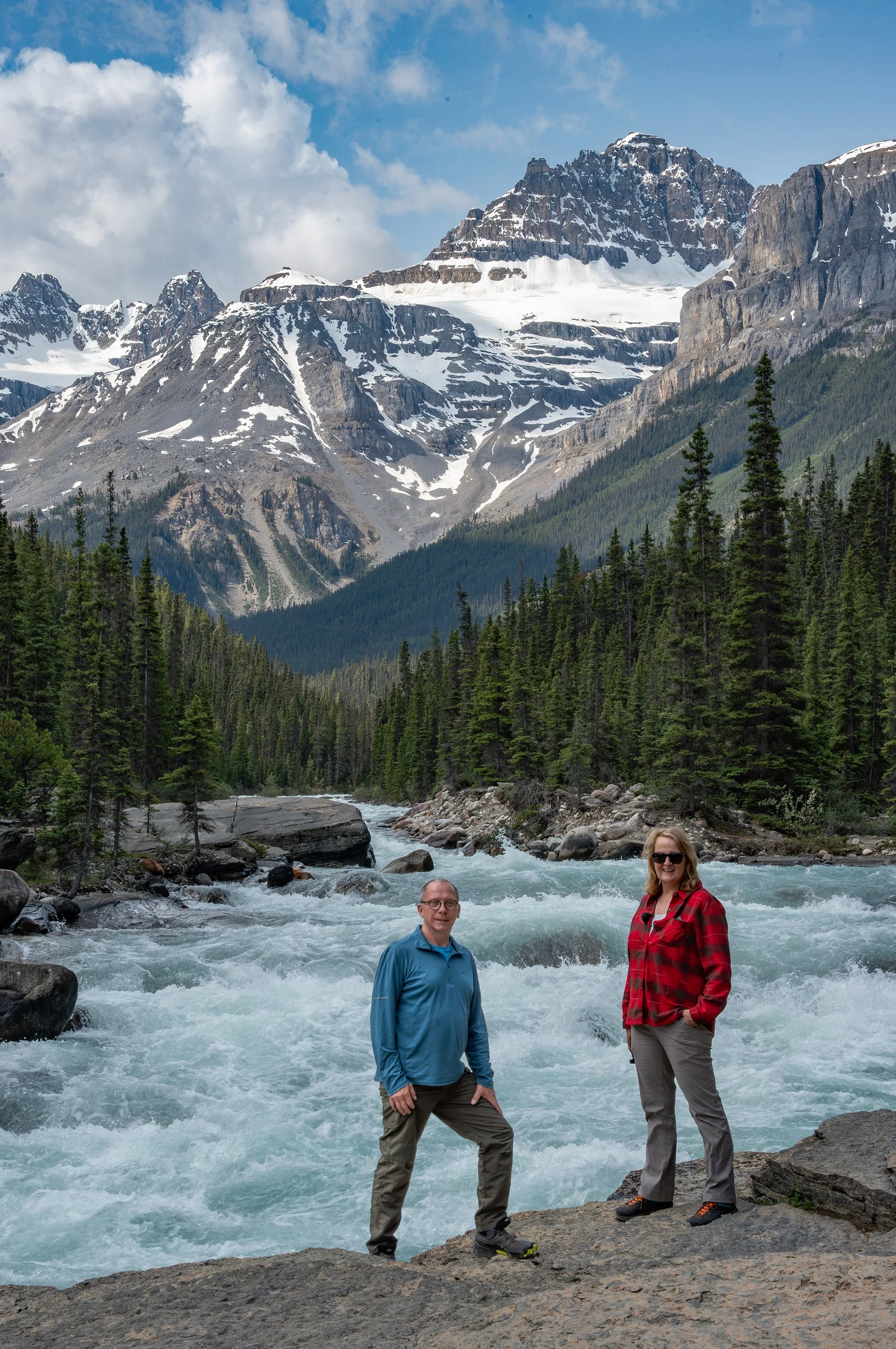 Two people stand on rocks in front of a rushing river with a forested mountain landscape and snow-capped peaks in the background.