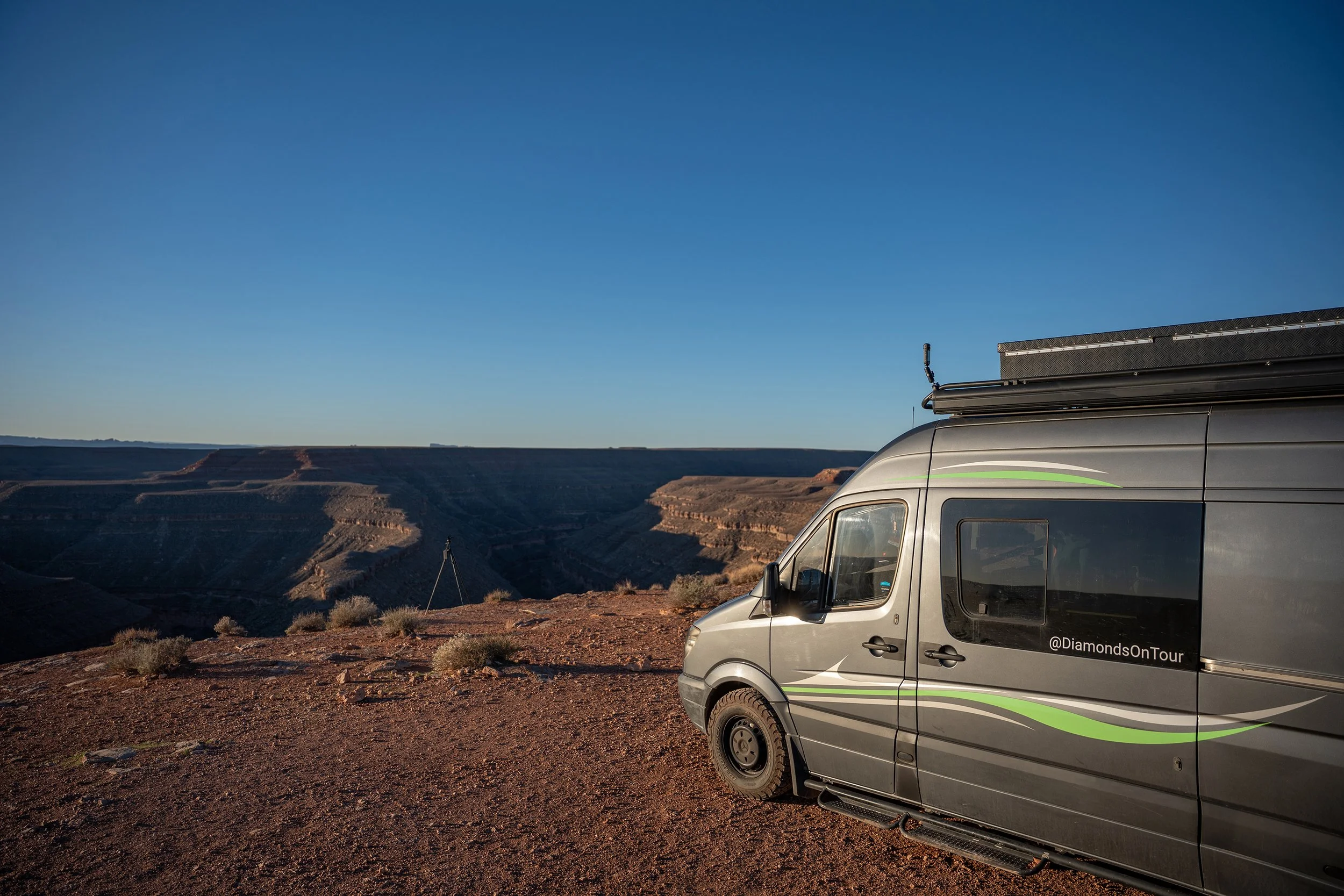 A silver camper van parked on a dirt ledge overlooking a canyon landscape under a clear blue sky. Goosenecks State park.