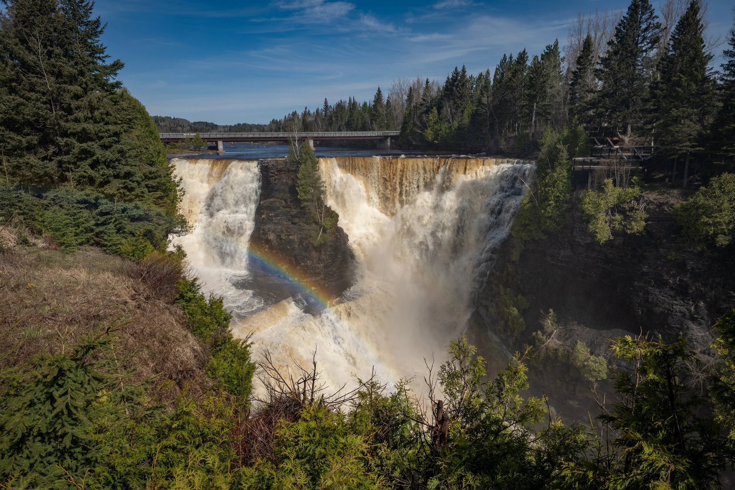 Waterfall surrounded by pine trees with a rainbow at the base and a bridge across the top under a blue sky with some clouds.