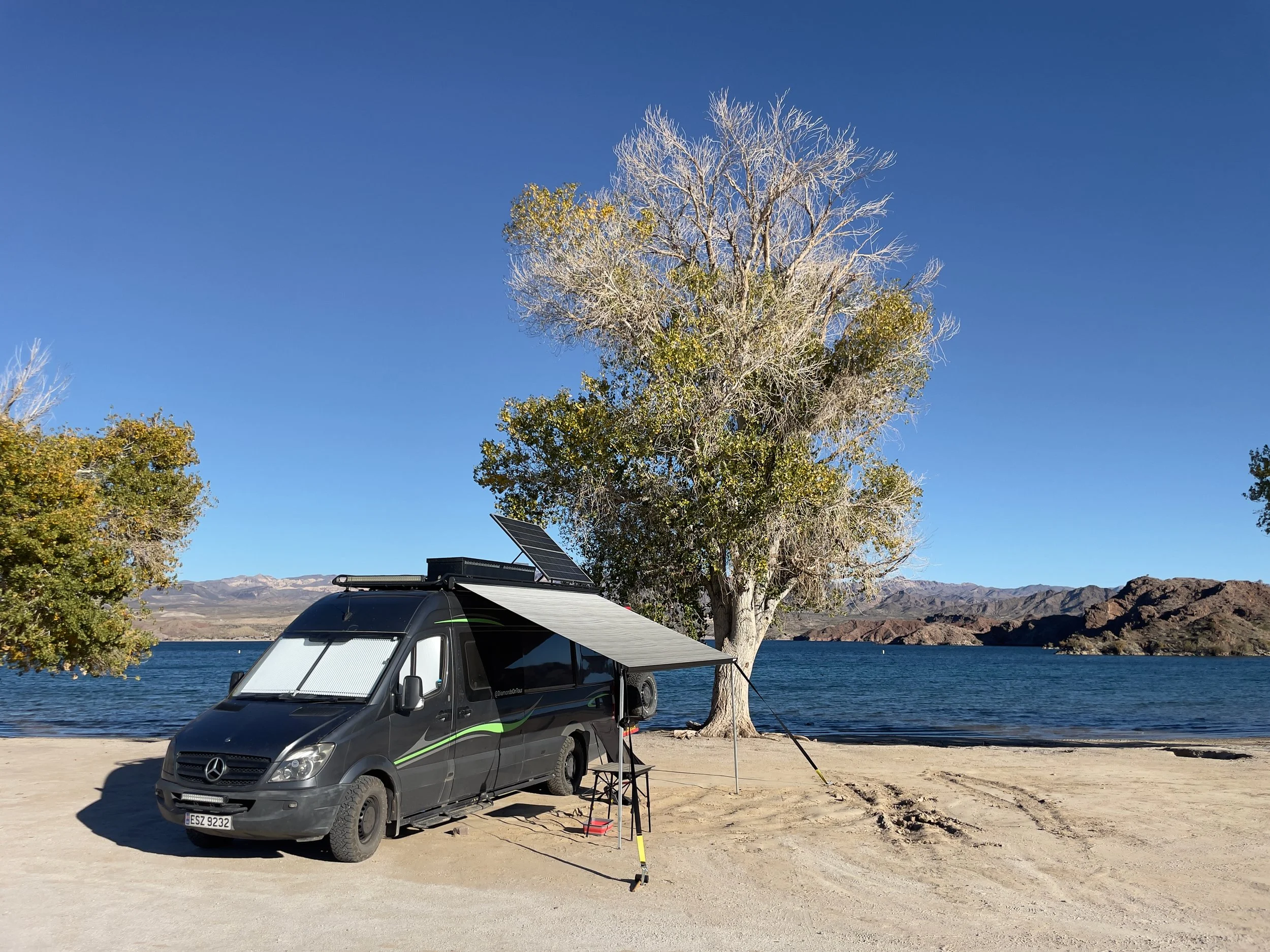 A dark gray camper van with green accents is parked at the edge of a lake, with solar panels on the roof. It is attached to a power source on the ground. Nearby, there is a black folding chair and a small table. Behind the van, there are trees with s