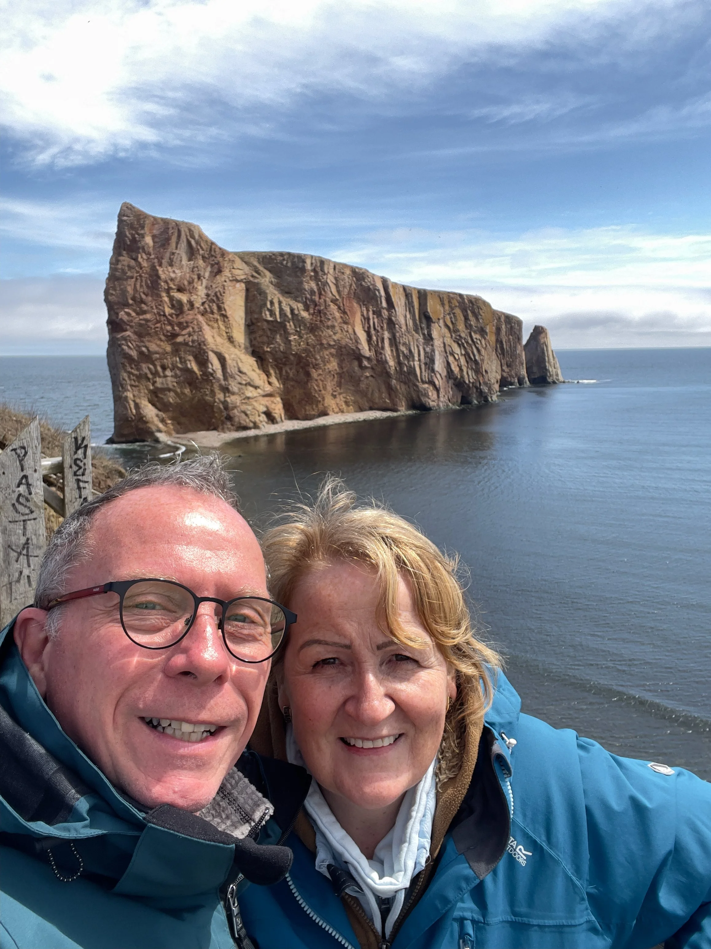 A selfie of two smiling older adults, a man and a woman, outdoors with a large rocky island and the ocean in the background. The man has glasses and gray hair, and the woman has curly blonde hair. They are dressed in outdoor jackets and appear to be 