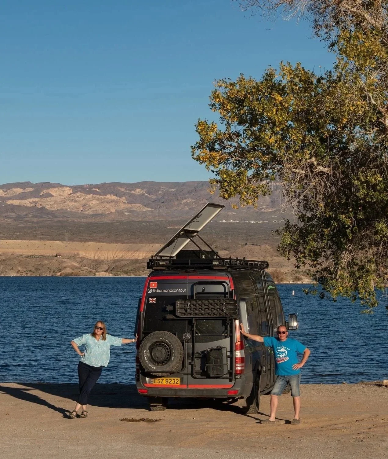 Two people standing next to a black van by a lake with mountains in the background. One woman on the left and one man on the right, posing with the van, which has a roof-mounted solar panel and various equipment. A large tree is on the right side of the image.