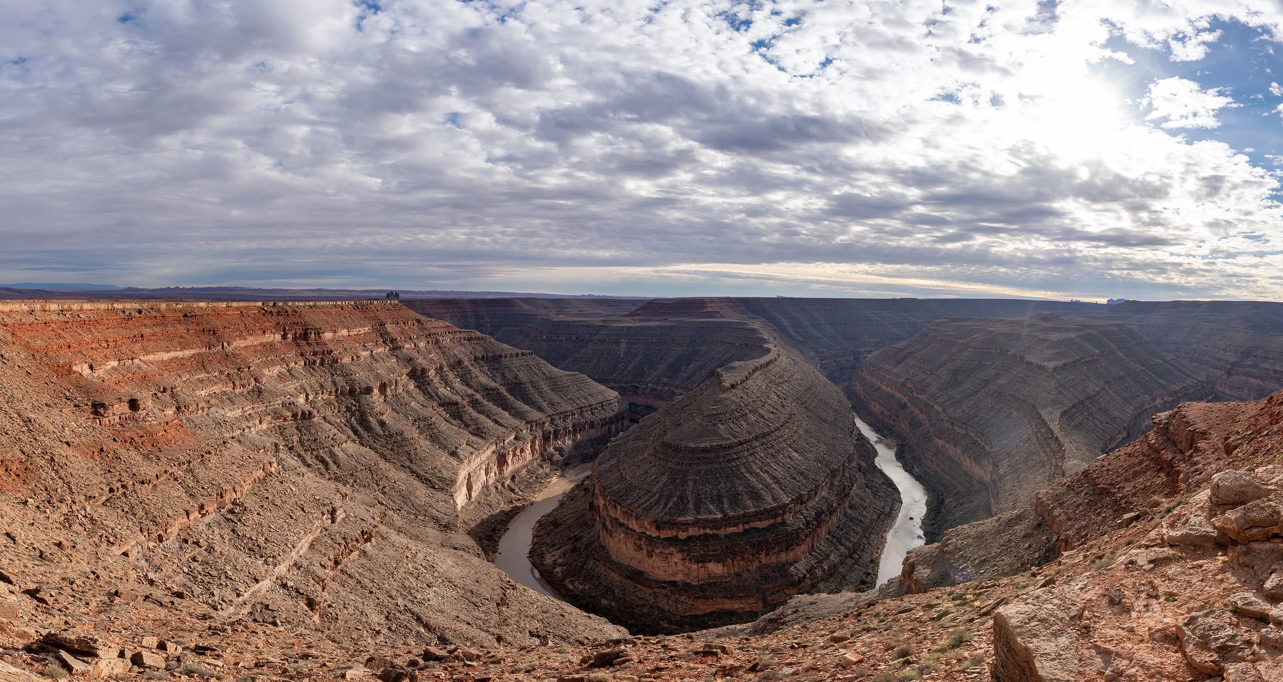 View of the Goosenecks State Park with layered rock formations and a river at the bottom under a partly cloudy sky.