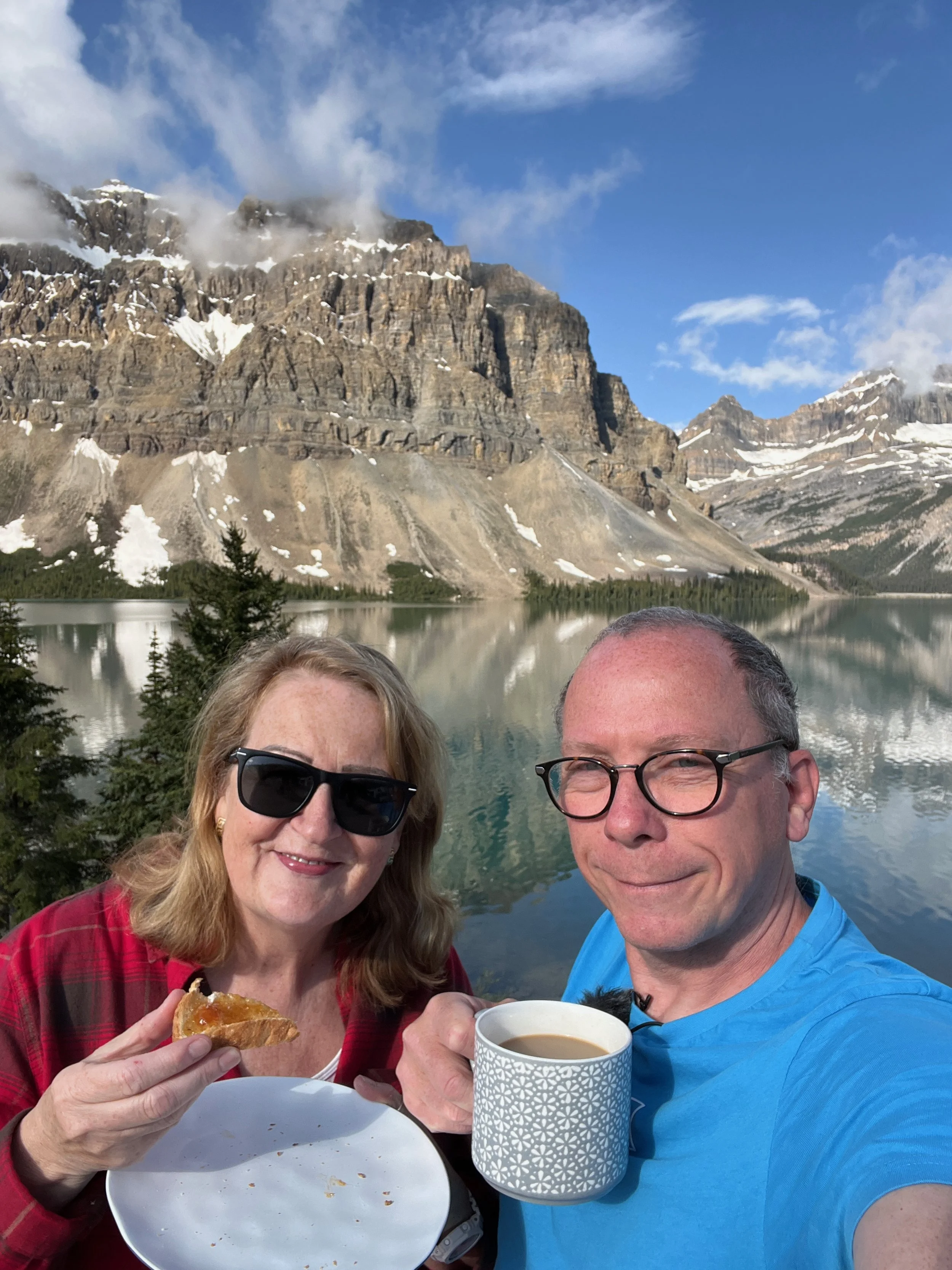 Two people enjoying breakfast outdoors with a mountain and lake in the background, one woman holding a piece of bread and the man holding a cup of coffee.