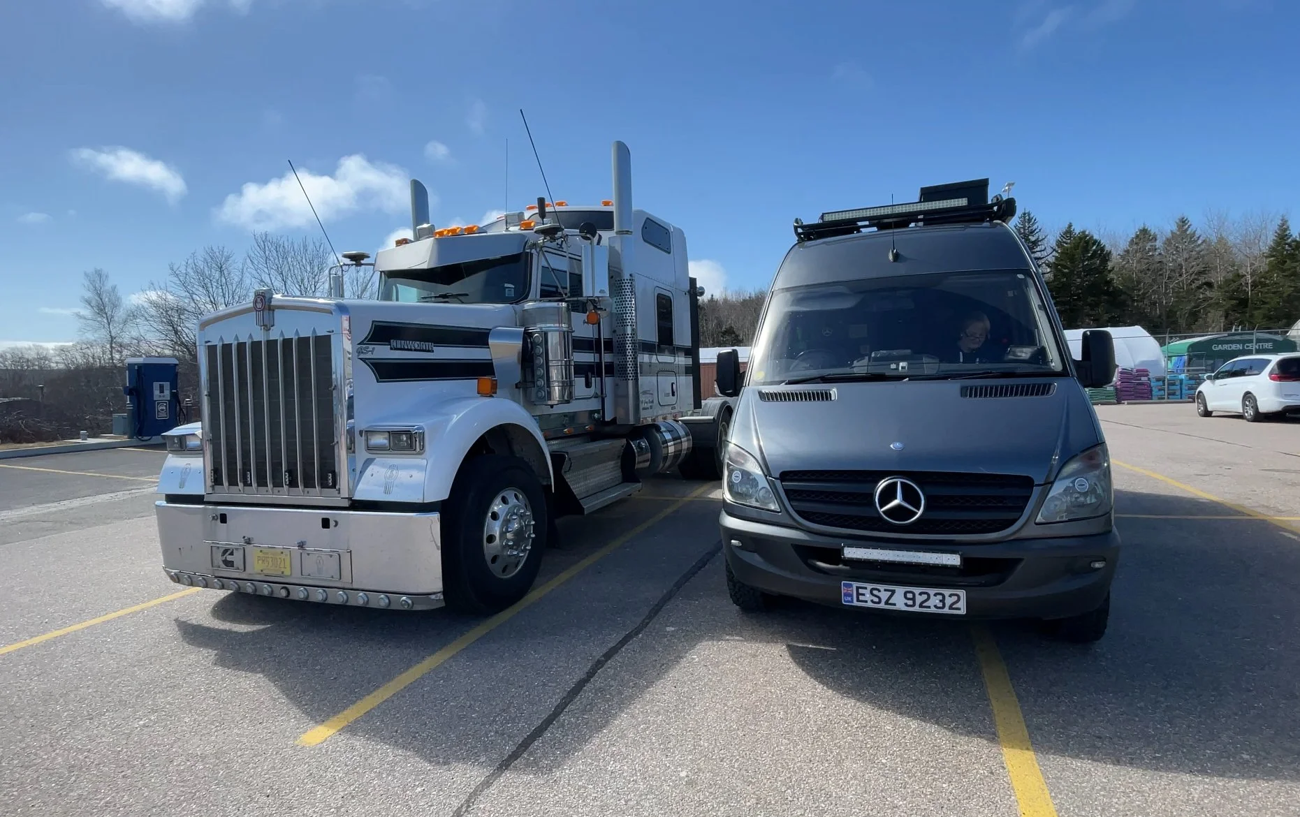 A parking lot with a white semi-truck and a black Mercedes-Benz van parked side by side on a sunny day. The semi-truck has a chrome grille, and the van has a roof rack. There are other cars and a garden center in the background, along with trees and 