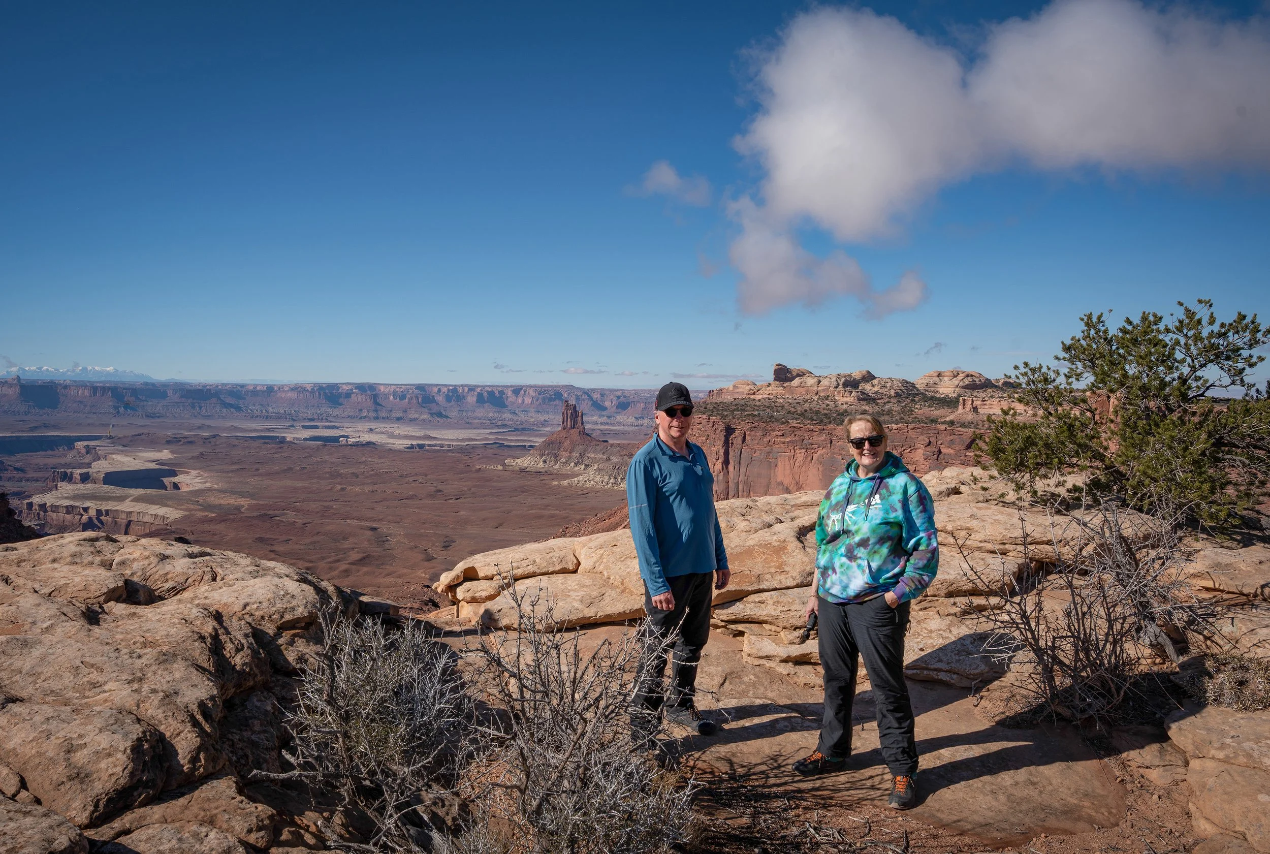Two people stand on a rocky ledge in a desert landscape with canyons and mesas, under a blue sky with scattered clouds.
