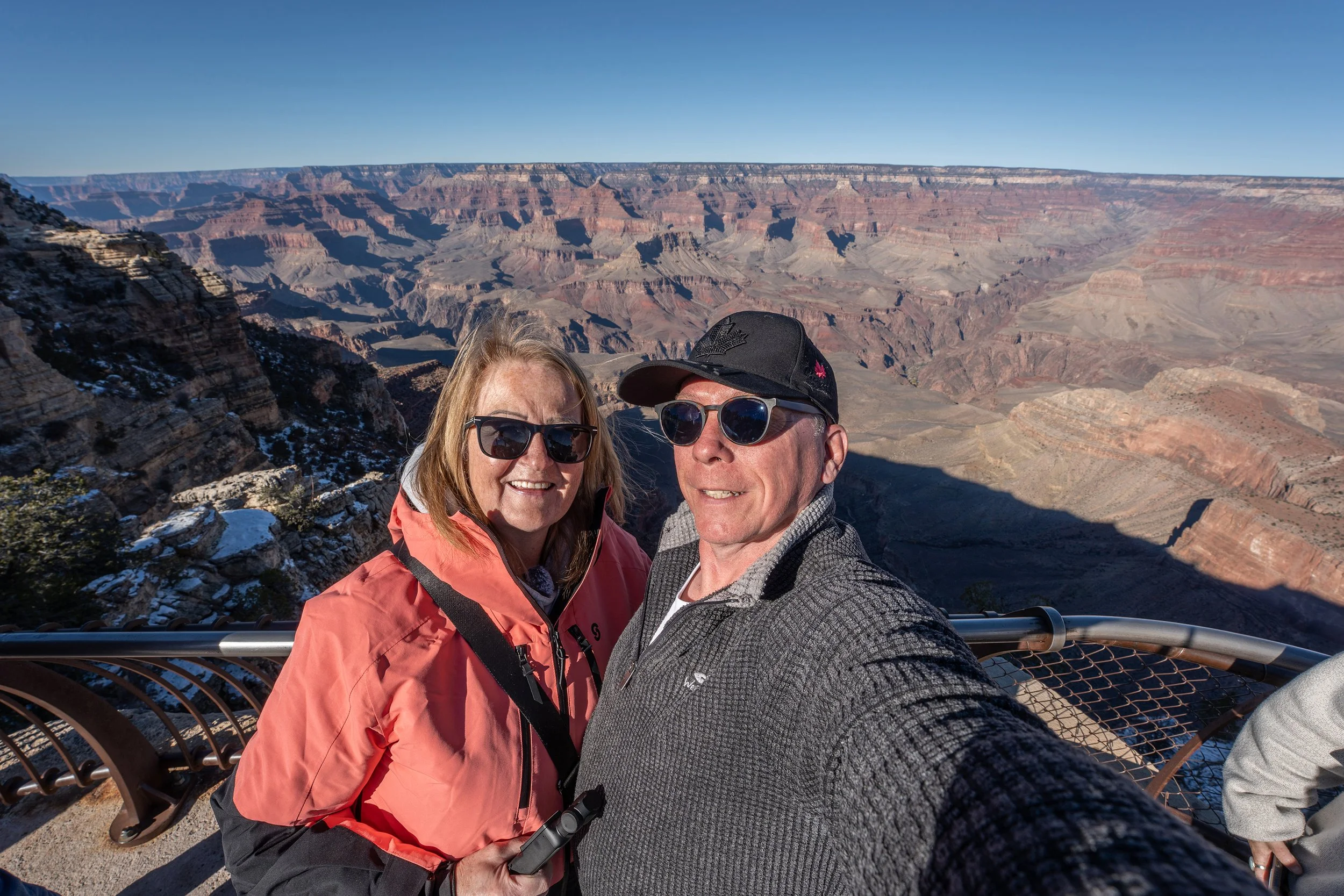 A smiling middle-aged man and woman taking a selfie at the Grand Canyon with the canyon's layered cliffs in the background, the woman holding a selfie stick and wearing sunglasses and an orange jacket, and the man wearing sunglasses, a black cap, and