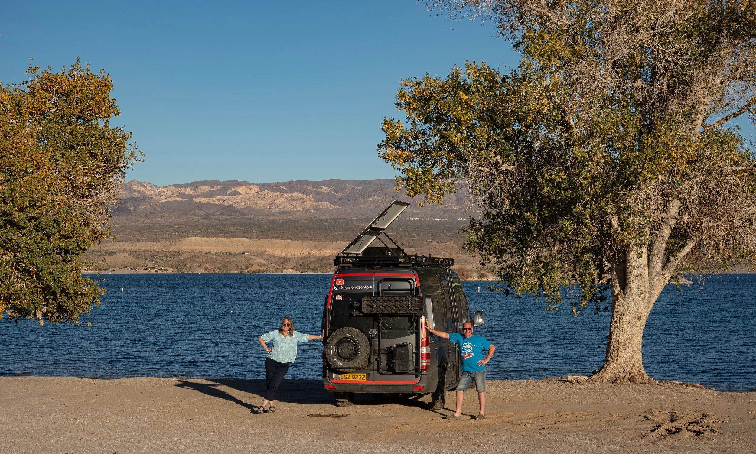 Two people standing next to a grey camper van with a solar panel on top, by a lake with trees and mountains in the background.