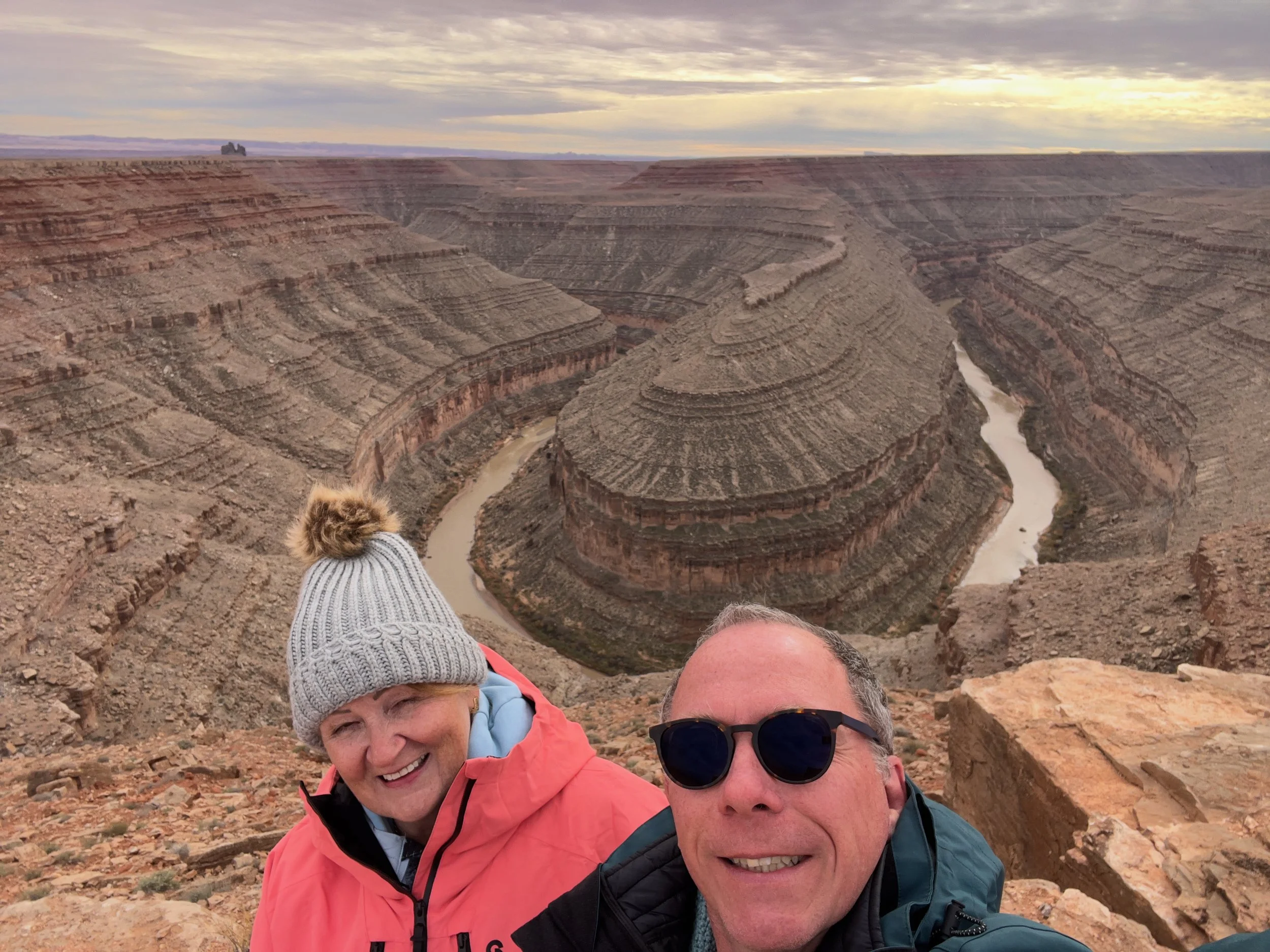A selfie of three people, two women and one man, at the Goosenecks State Park with a vast canyon view and the Colorado River in the background, under cloudy sky.
