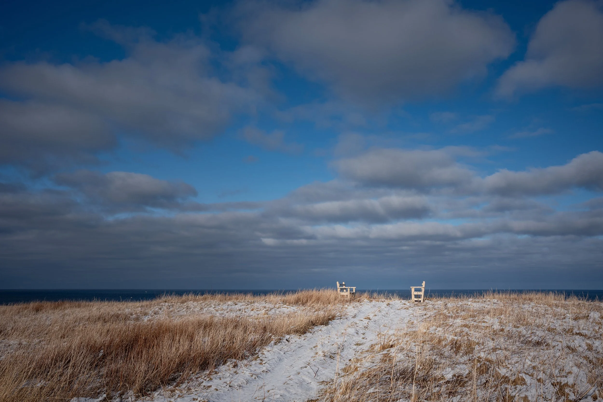 A beach with sandy and grassy shore, two benches facing the ocean, cloudy sky above, and calm water in the background.