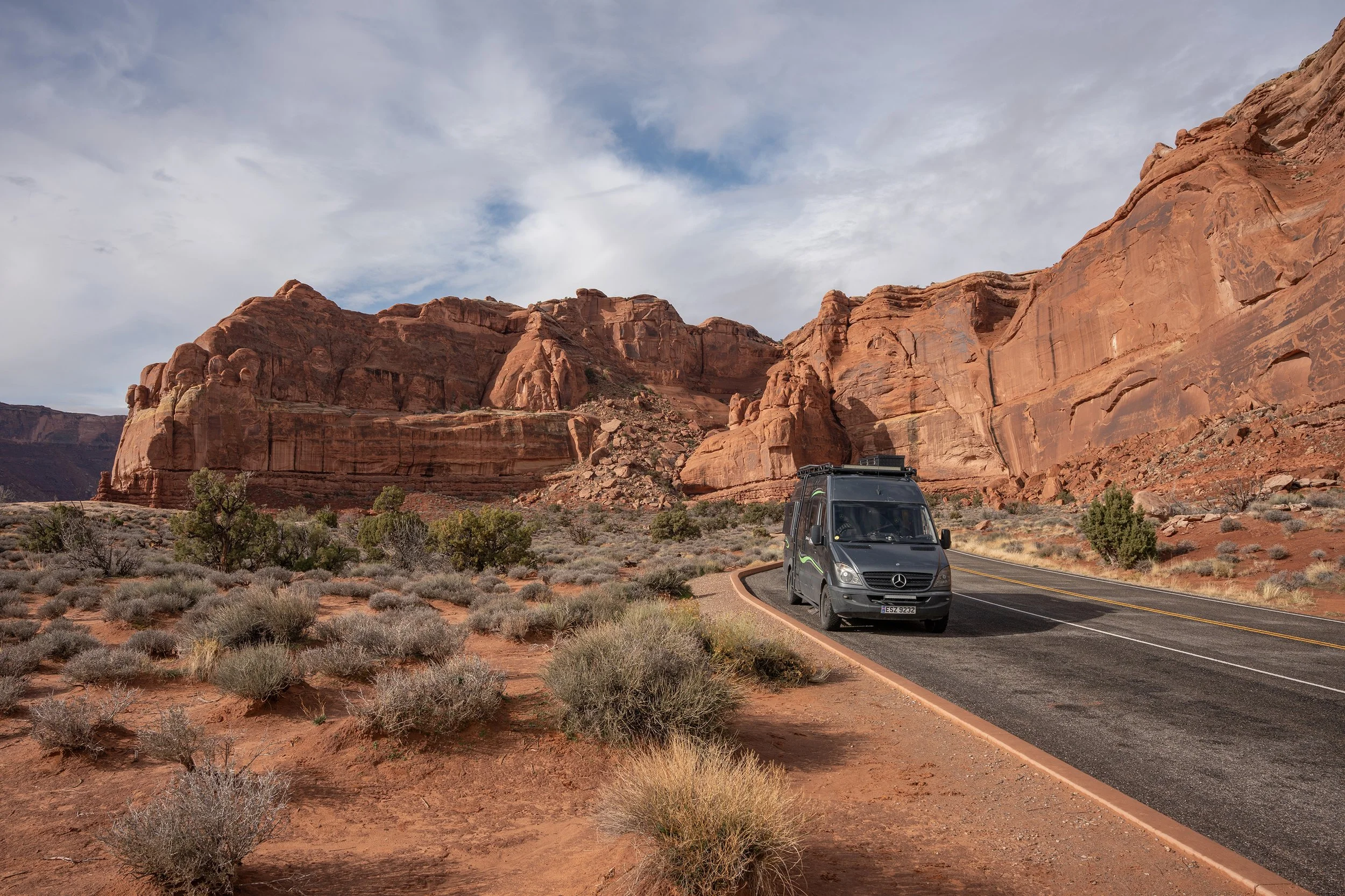 A black Mercedes-Benz van parked on a winding road in a desert landscape with large red rock formations and sparse desert vegetation under a partly cloudy sky.