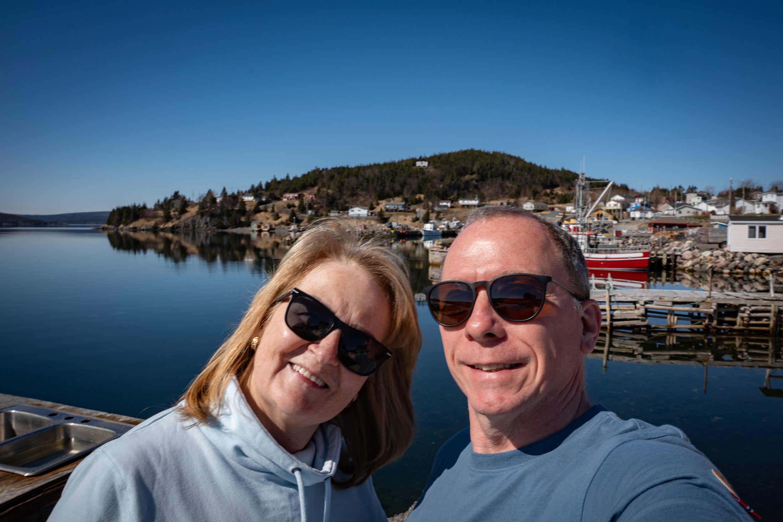 Two people wearing sunglasses taking a selfie by the water, with a hillside and houses in the background. Dildo, Newfoundland, Canada,