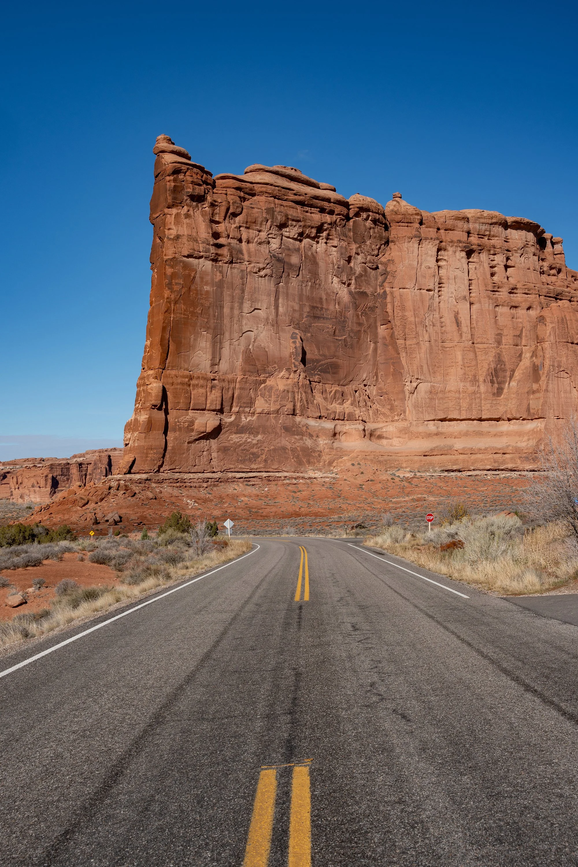 A paved road with yellow dividing lines, leading towards a large red rock formation in a desert landscape, with clear blue sky overhead.