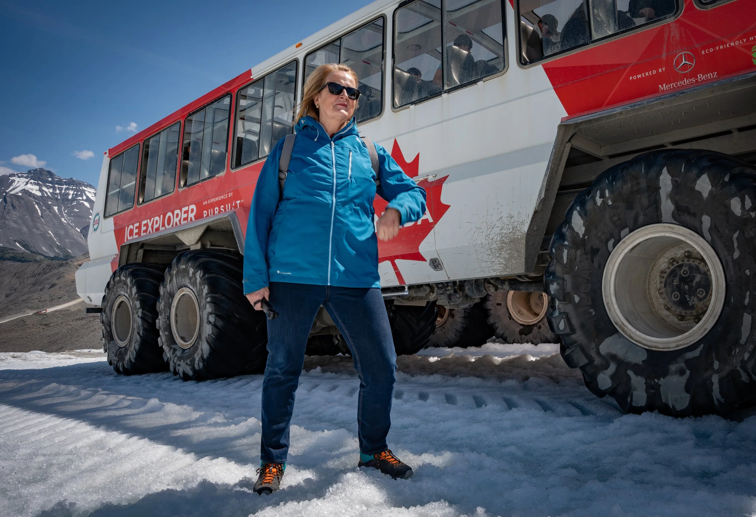 A woman wearing sunglasses, a blue jacket, and hiking shoes standing on snow-covered ground in front of a large red and white tracked vehicle labeled 'Ice Explorer' with mountains in the background.