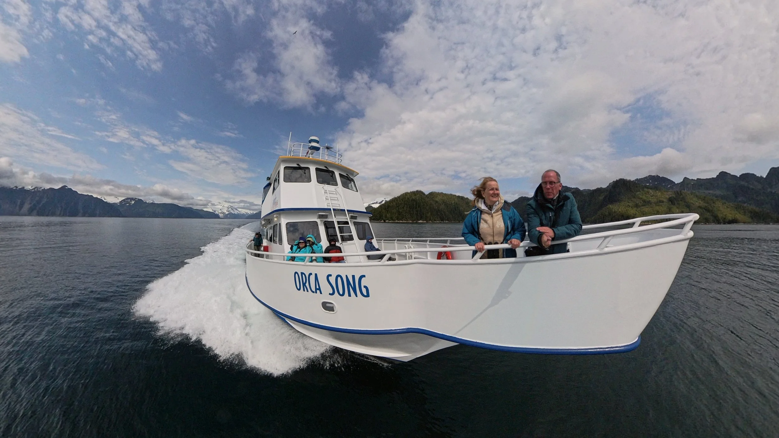 A white boat labeled 'ORCA SONG' on water with two people standing at the front, smiling, and surrounded by a scenic landscape of mountains and cloudy sky. Alaska, USA.
