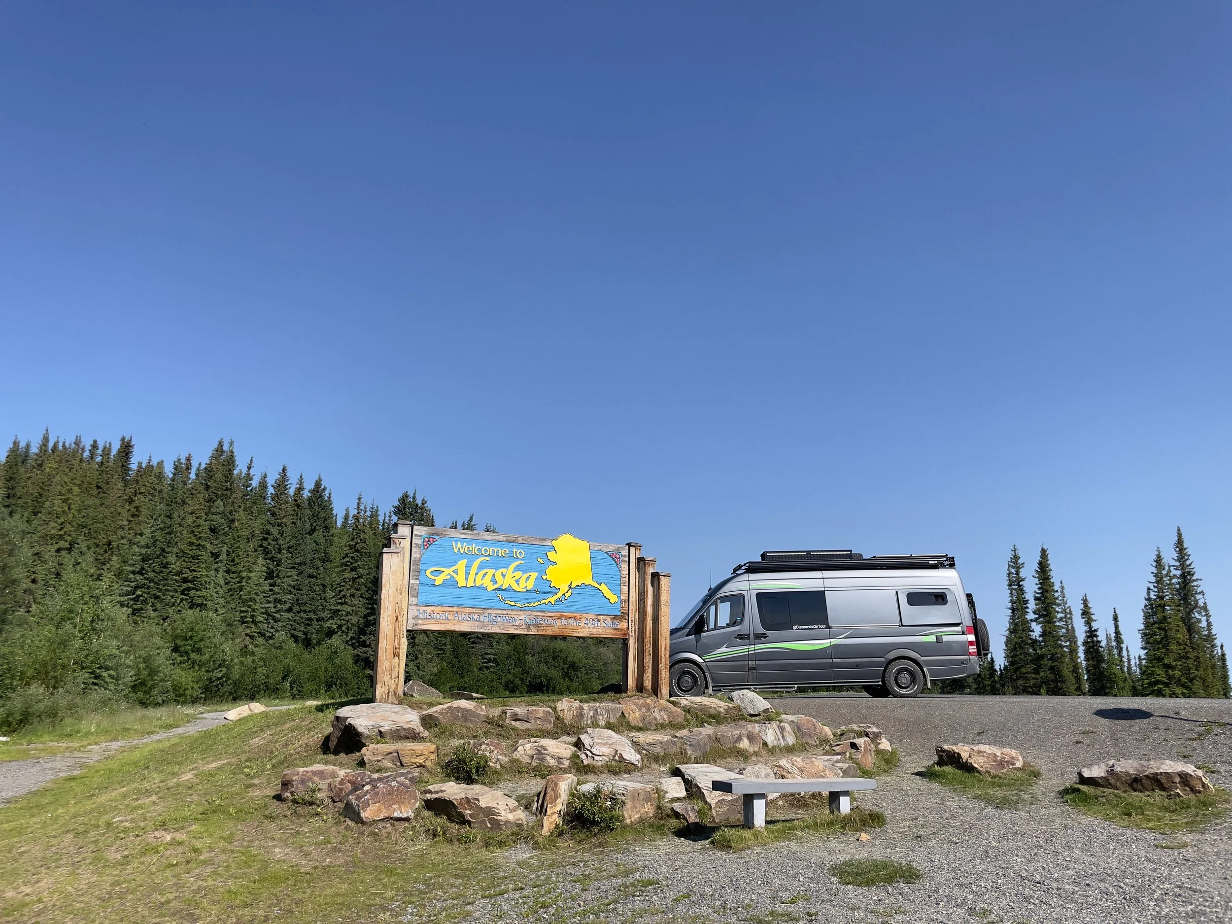 A scenic outdoor view of a "Welcome to Alaska" sign on a wooden board with a map of Alaska, surrounded by rocks and grass, with a grey camper van parked to the right, and dense green trees in the background under a clear blue sky.