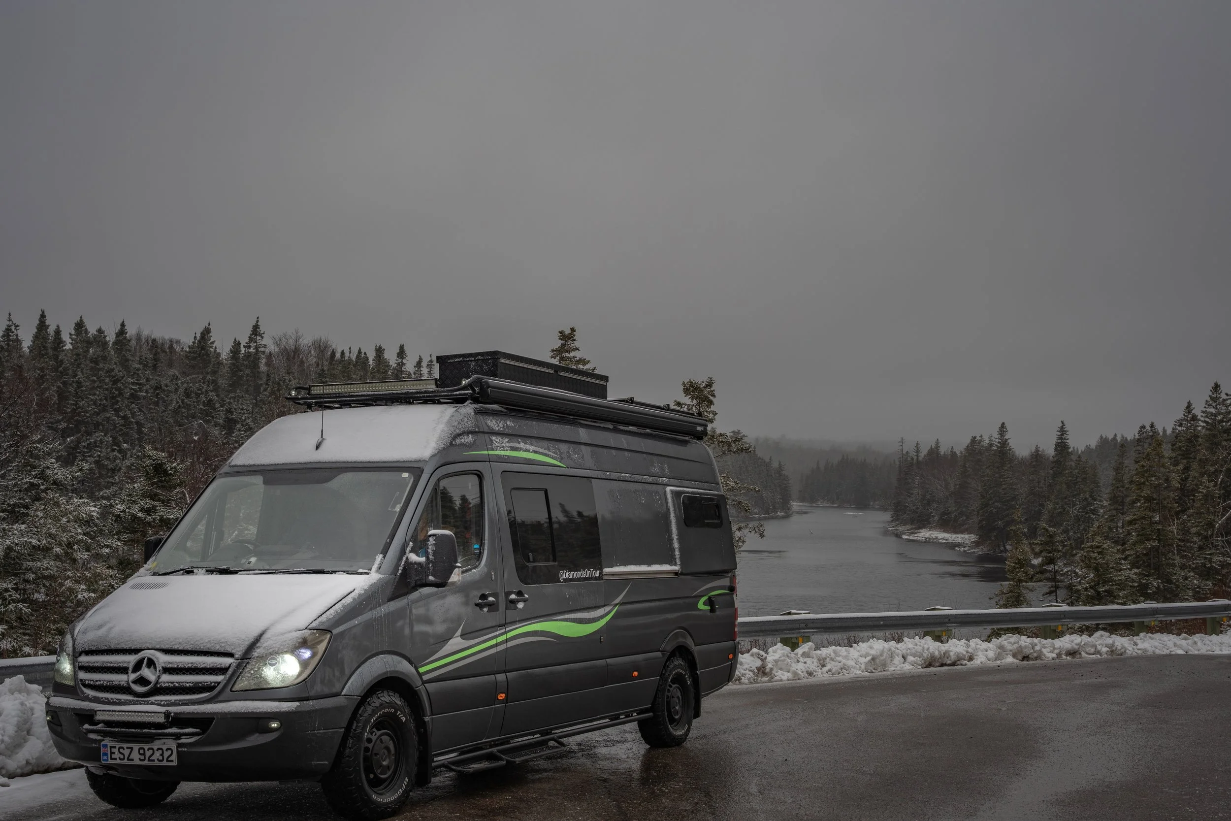 A dark gray Mercedes-Benz camper van parked on a snowy roadside near a river, with snow-covered trees and an overcast sky in the background.