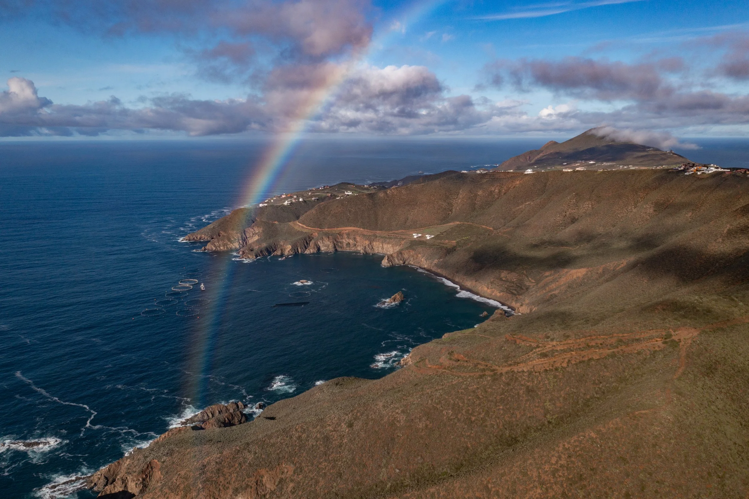 A scenic view of the ocean coastline with rugged cliffs, a rainbow, and clouds over a mountain in the distance.