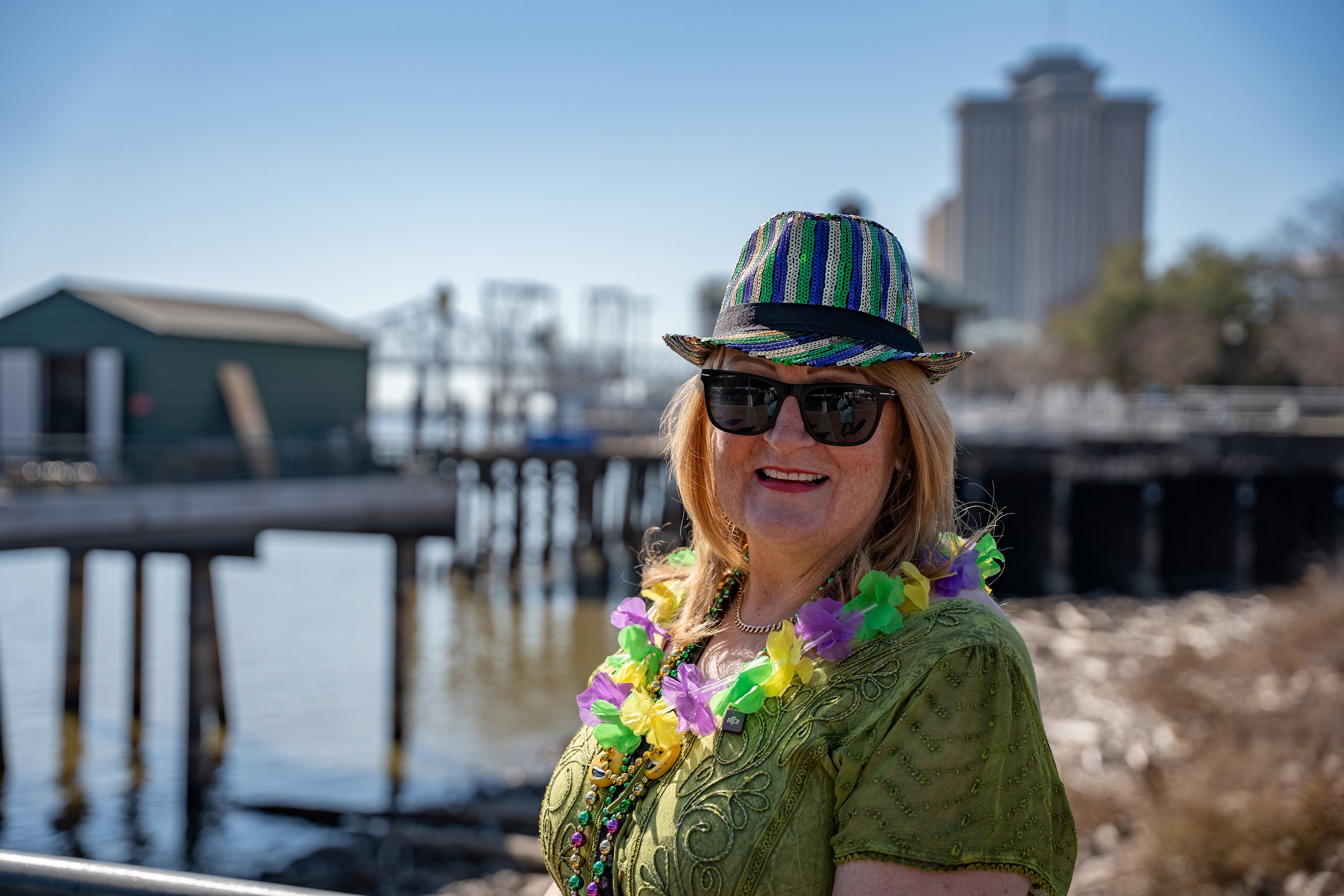A woman with light skin and red hair wearing sunglasses, a colorful beaded hat, flower leis, and a green embroidered shirt, smiling at a waterfront with a pier and large building in the background.