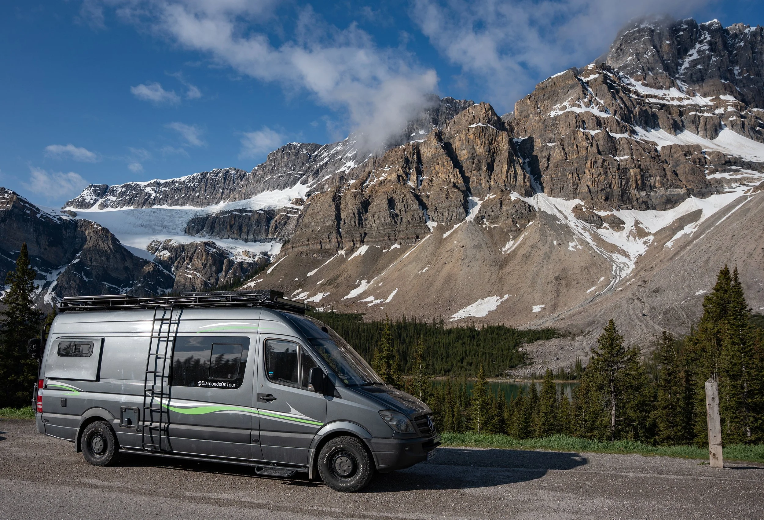 A gray camper van parked in front of a mountain range with snow, trees, and a partly cloudy blue sky. Rocky Mountains, Canada.