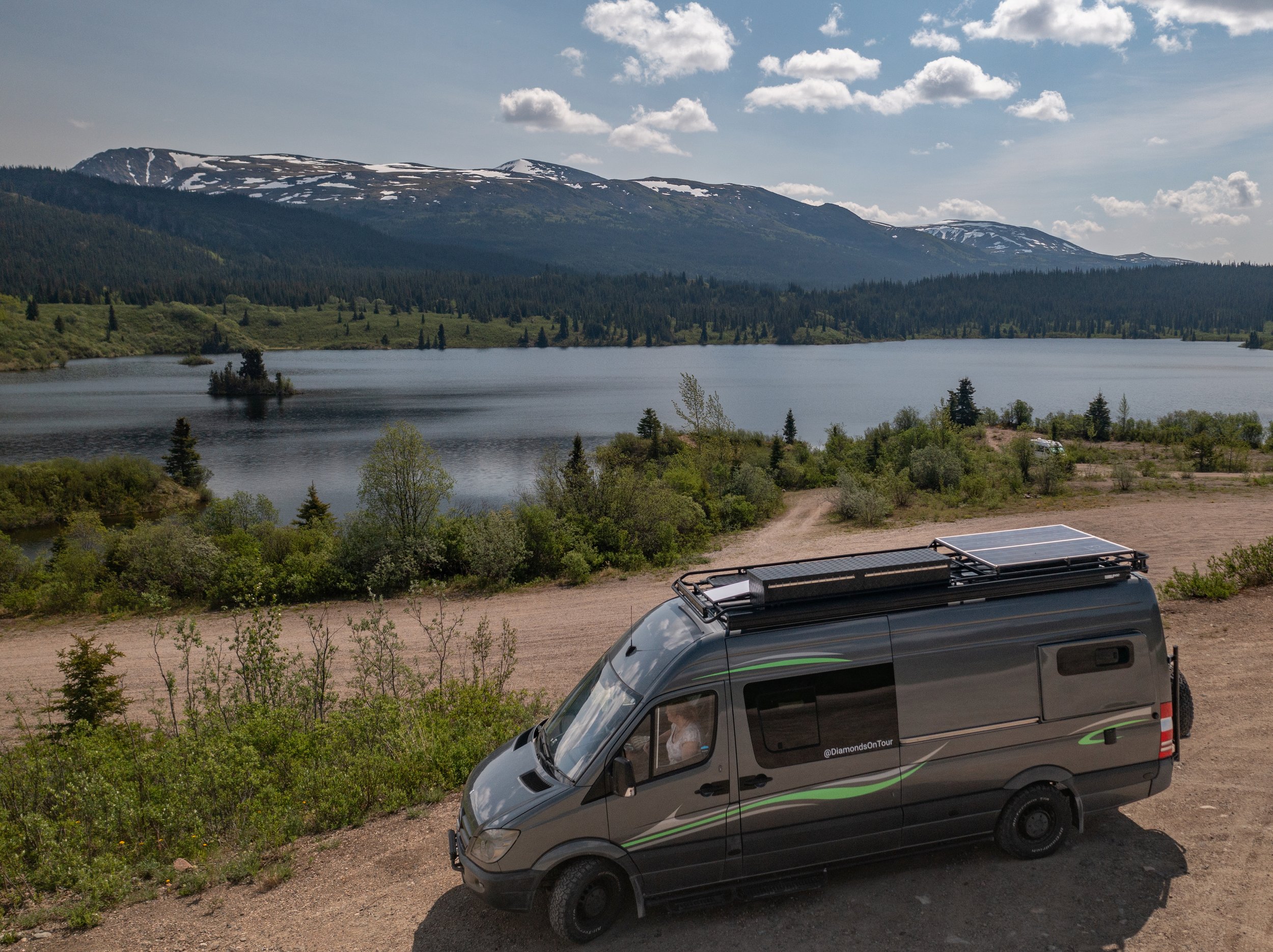 A scenic view of a lake with mountains in the background, partially covered with snow, under a partly cloudy sky. In the foreground, a black van with green accents and a roof rack is parked, with some greenery and dirt path around it.