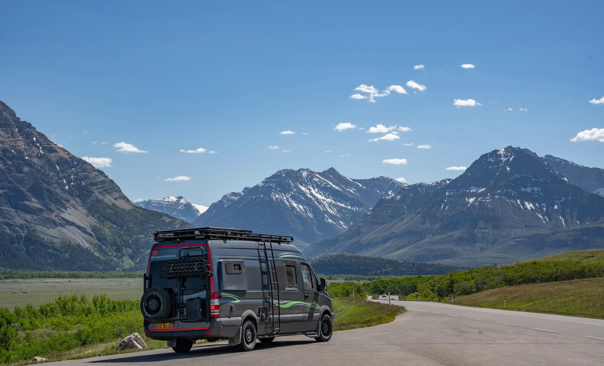 A van parked on the side of a road in front of a scenic mountain landscape with snow-capped peaks and a blue sky with scattered clouds. Dempster Highway, Yukon Territory, Canada.