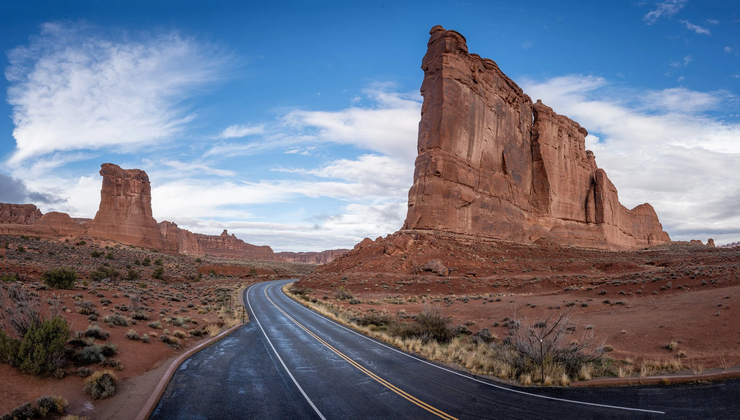 A winding road through a desert landscape with large rock formations under a blue sky with clouds