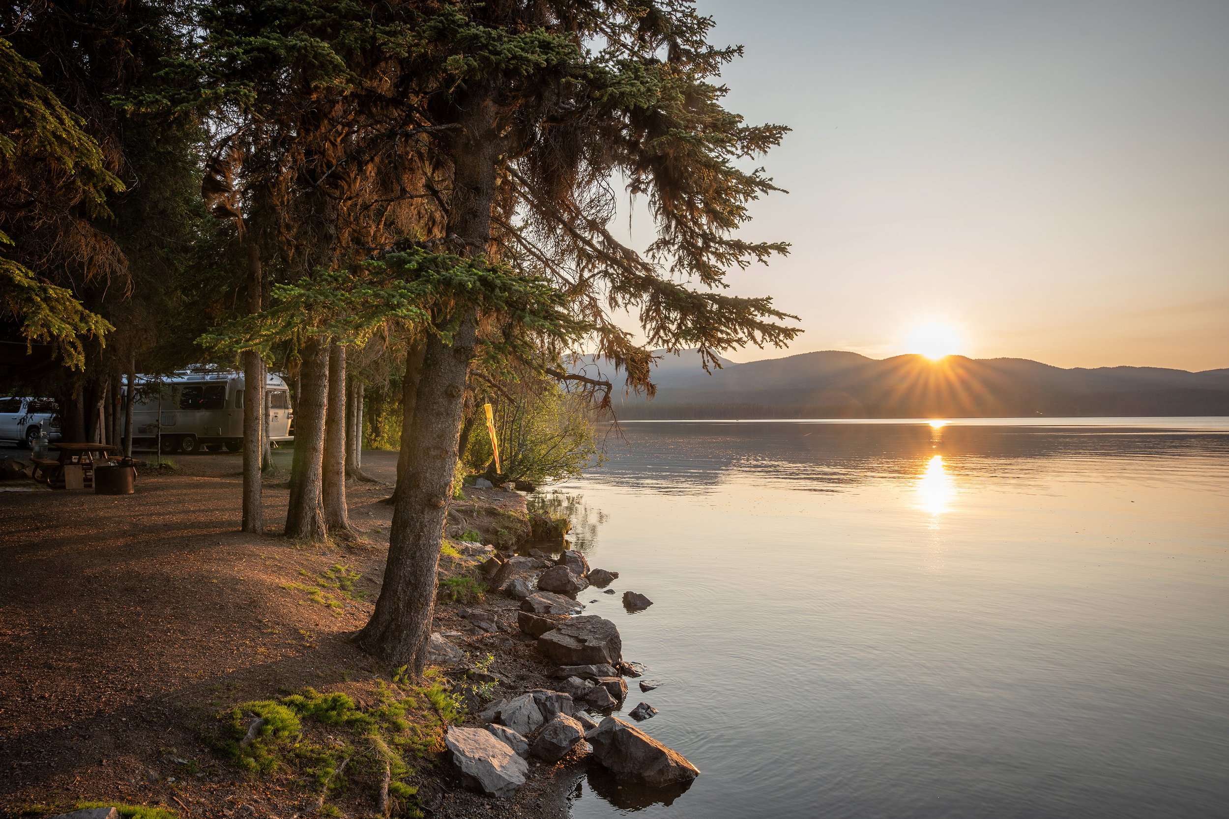 Sunset over a lake with mountains in the background, dense trees on the left, and a rocky shoreline.